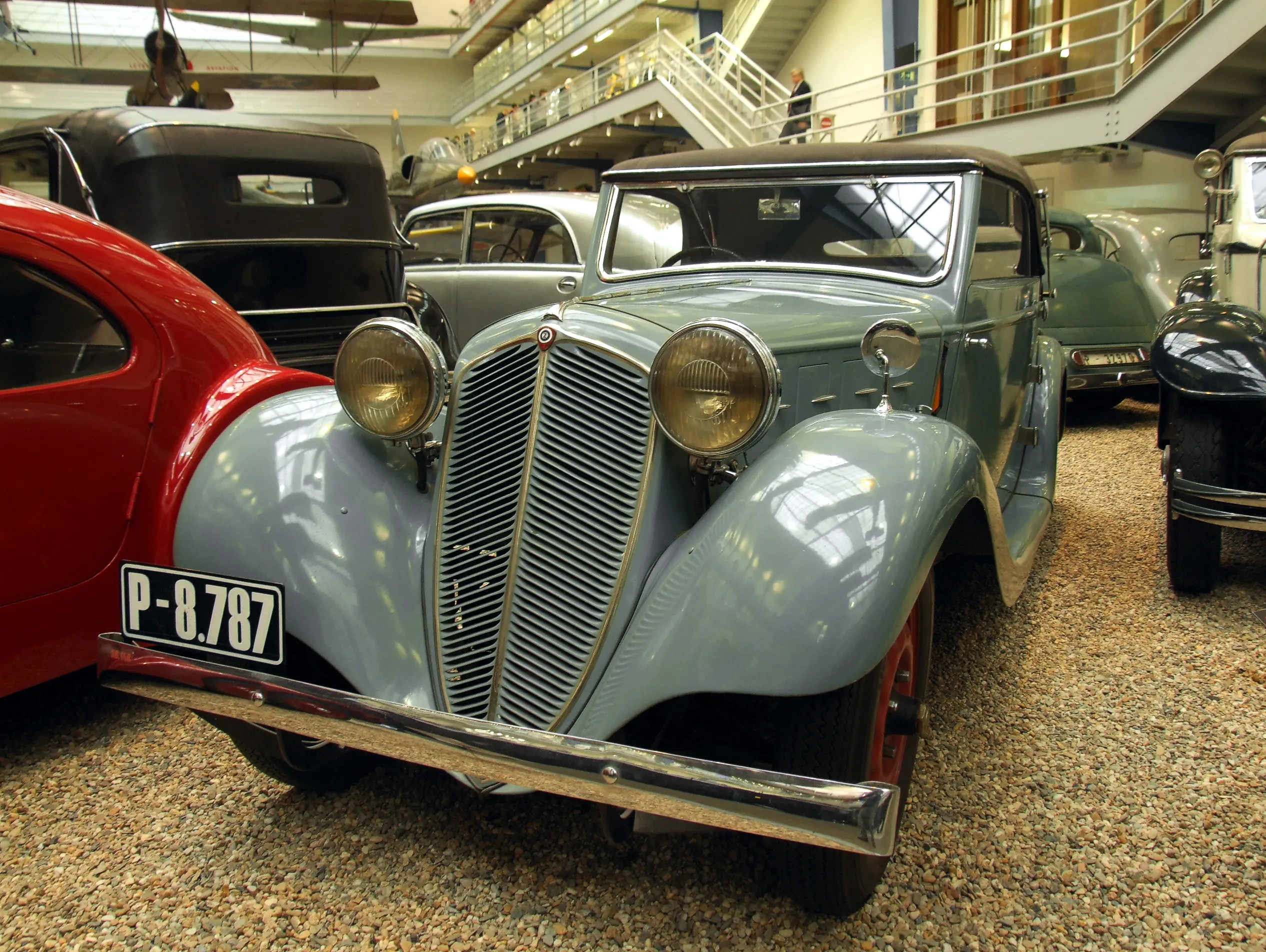 A light blue vintage car with a prominent grille and round headlights, parked indoors among other classic vehicles