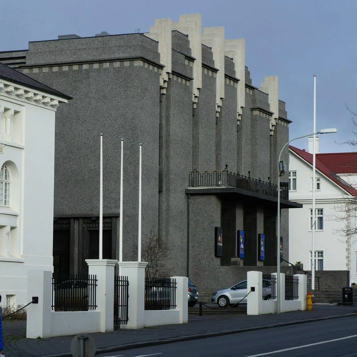 Buitenaanzicht van het National Theatre of Iceland, een grijs gebouw in art-decostijl met verticale betonnen vinnen langs de daklijn, witte vlaggenmasten en een zwart ijzeren hek.