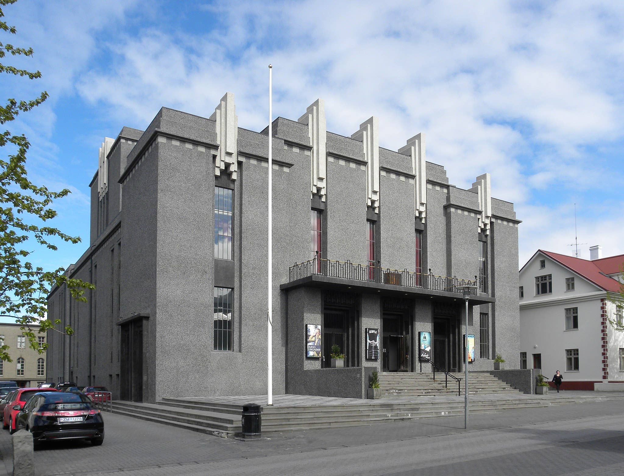 Grijs betonnen gebouw met verticale architectonische vinnen langs de gevel, trappen naar de ingang en geparkeerde auto's voor de deur