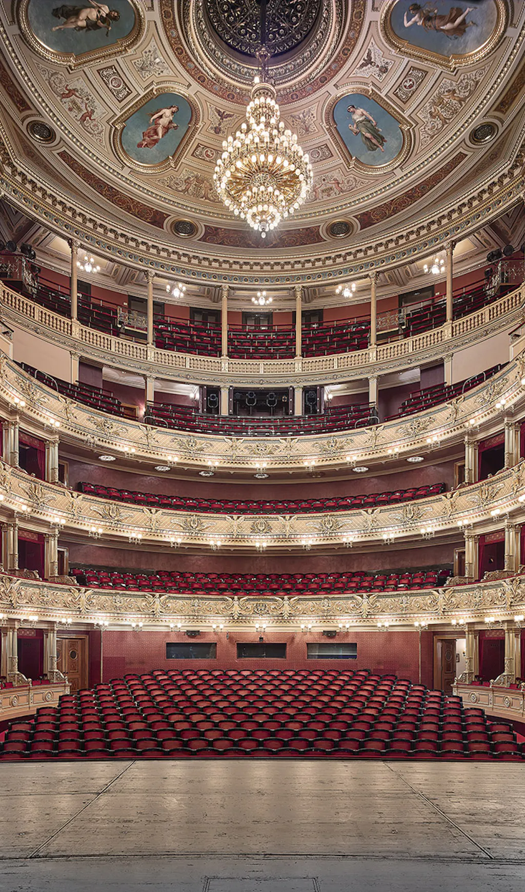 Sierlijk theaterinterieur met rode stoelen, meerdere balkons en een grote kroonluchter onder een versierd plafond