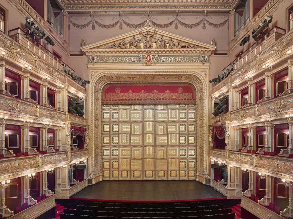 Ornate theater interior with multiple levels of balconies, red velvet seats, and a golden proscenium arch