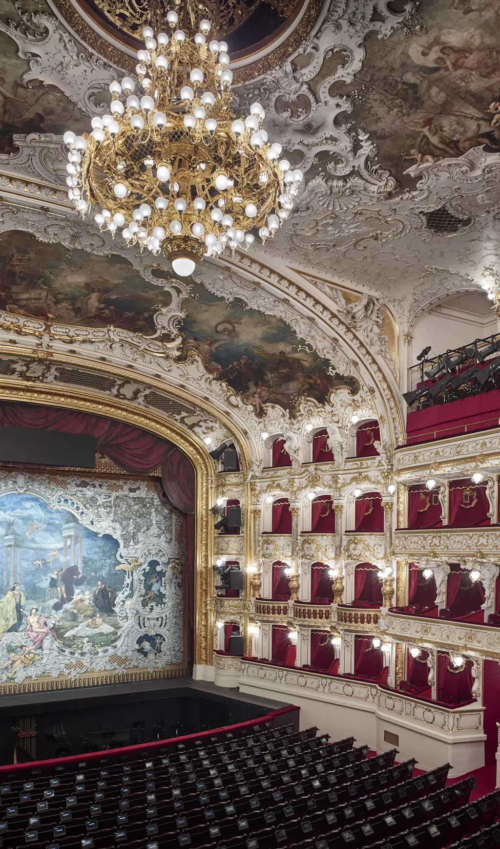 Ornate theater interior with a large chandelier, red seats, and decorative ceiling frescoes