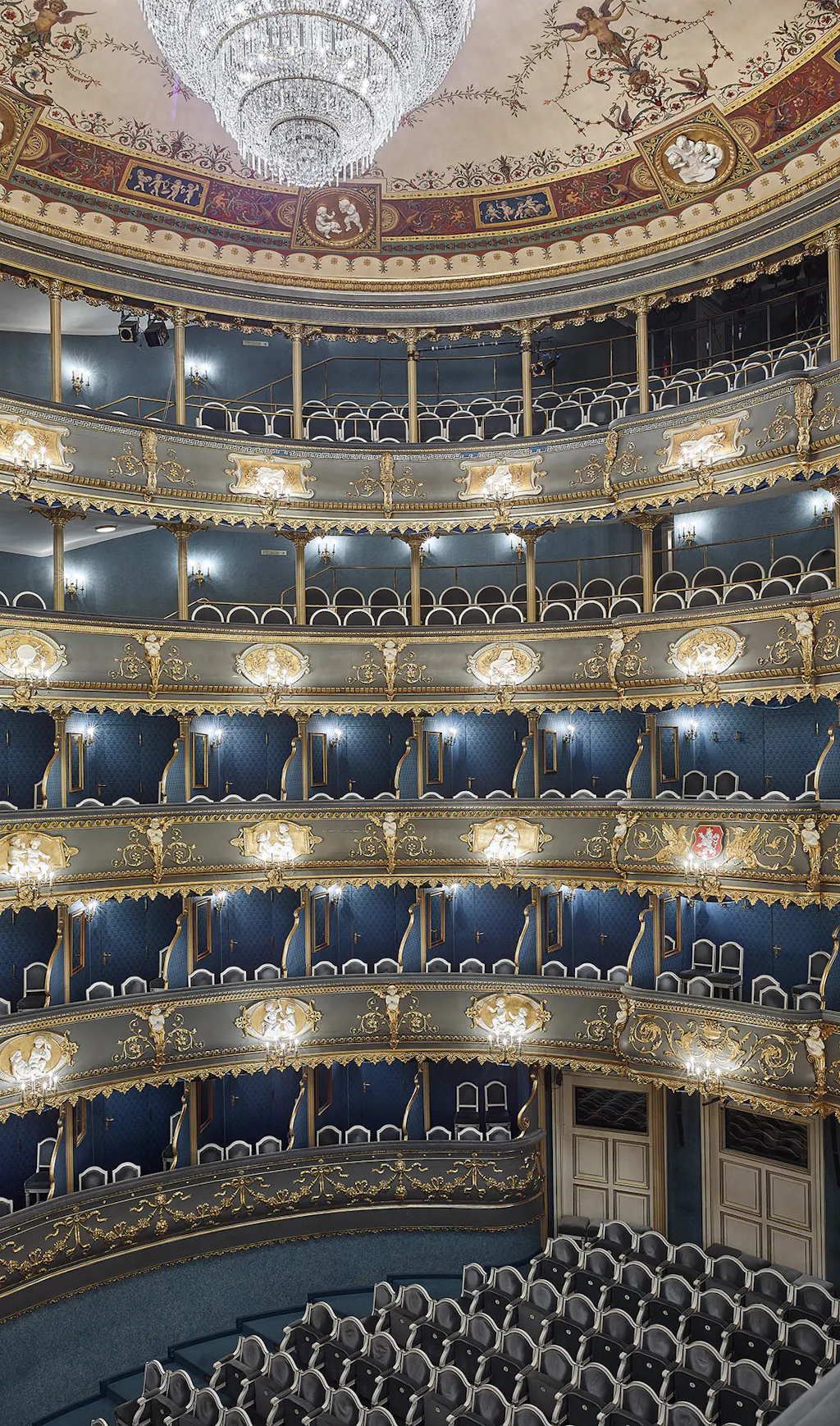 Ornate theater interior with multiple tiers of balconies, gilded decorations, blue seats, and a large crystal chandelier