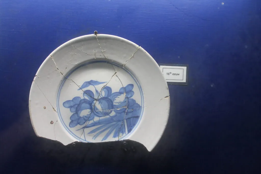 Plato de cerámica blanca roto con decoración floral azul expuesto sobre un fondo azul oscuro con una etiqueta pequeña