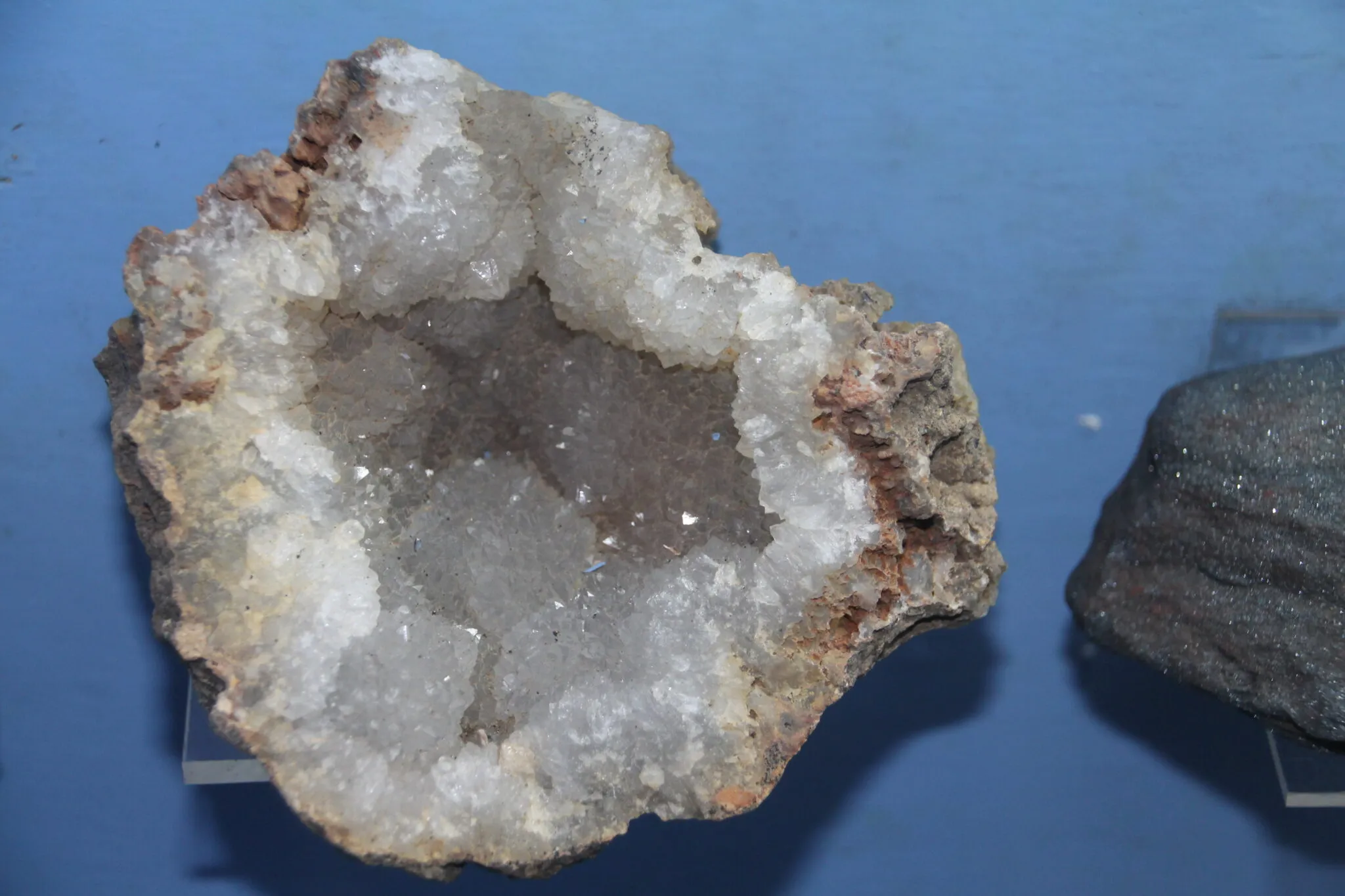 Una geoda con interior de cristales blancos y exterior rocoso, expuesta sobre un soporte transparente contra un fondo azul, con una roca oscura parcialmente visible a la derecha.