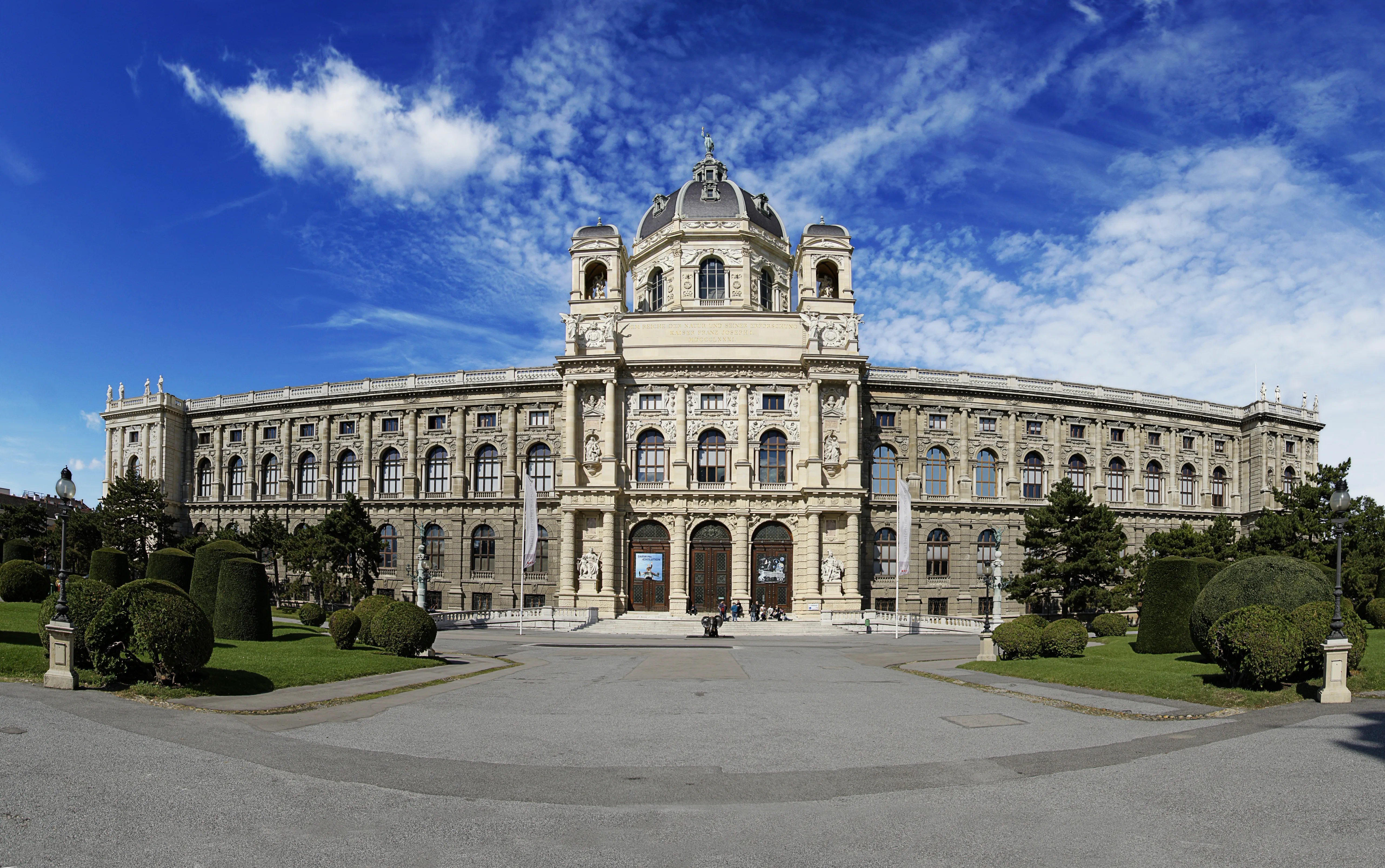 Muzeum Historii Naturalnej w Wiedniu z jego wielką neoklasycystyczną fasadą, centralną kopułą i symetrycznymi skrzydłami pod błękitnym niebem z białymi chmurami.