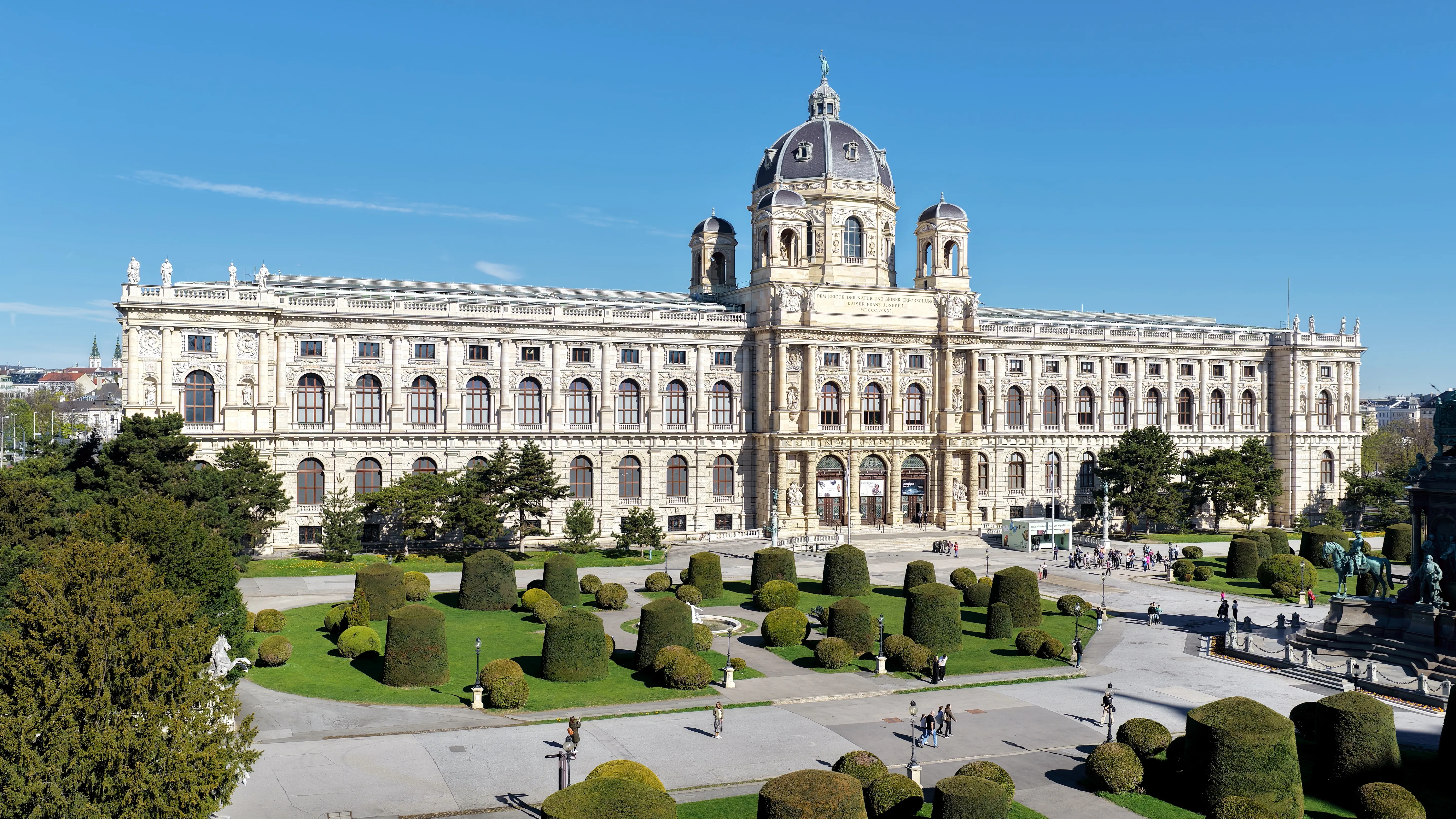 Szeroki widok na zewnętrzną część Muzeum Historii Naturalnej w Wiedniu, ukazujący okazałą neoklasycystyczną fasadę z centralną kopułą, łukowatymi oknami oraz zadbany ogród z przyciętymi okrągłymi krzewami pod bezchmurnym niebem.