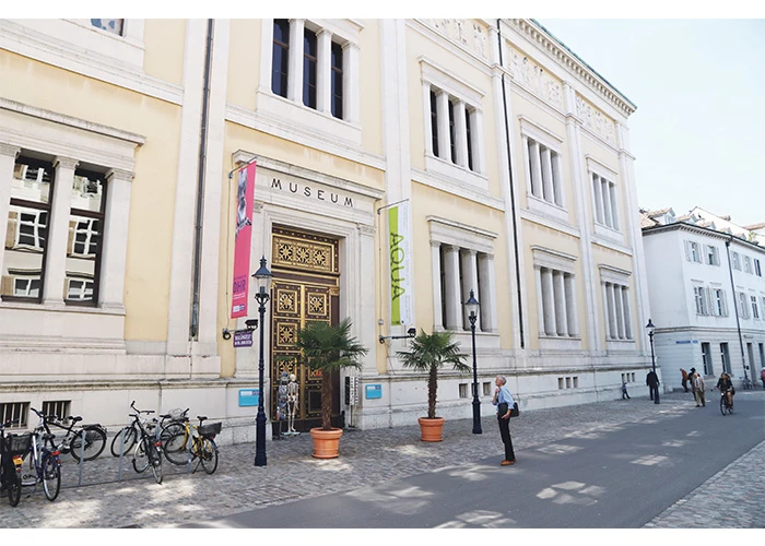 Exterieur van de gevel van het Naturhistorisches Museum Basel met de hoofdingang, het bordje MUSEUM, sierlijke gouden deuren, roze en groene banieren, palmbomen in potten en fietsen die buiten geparkeerd staan.