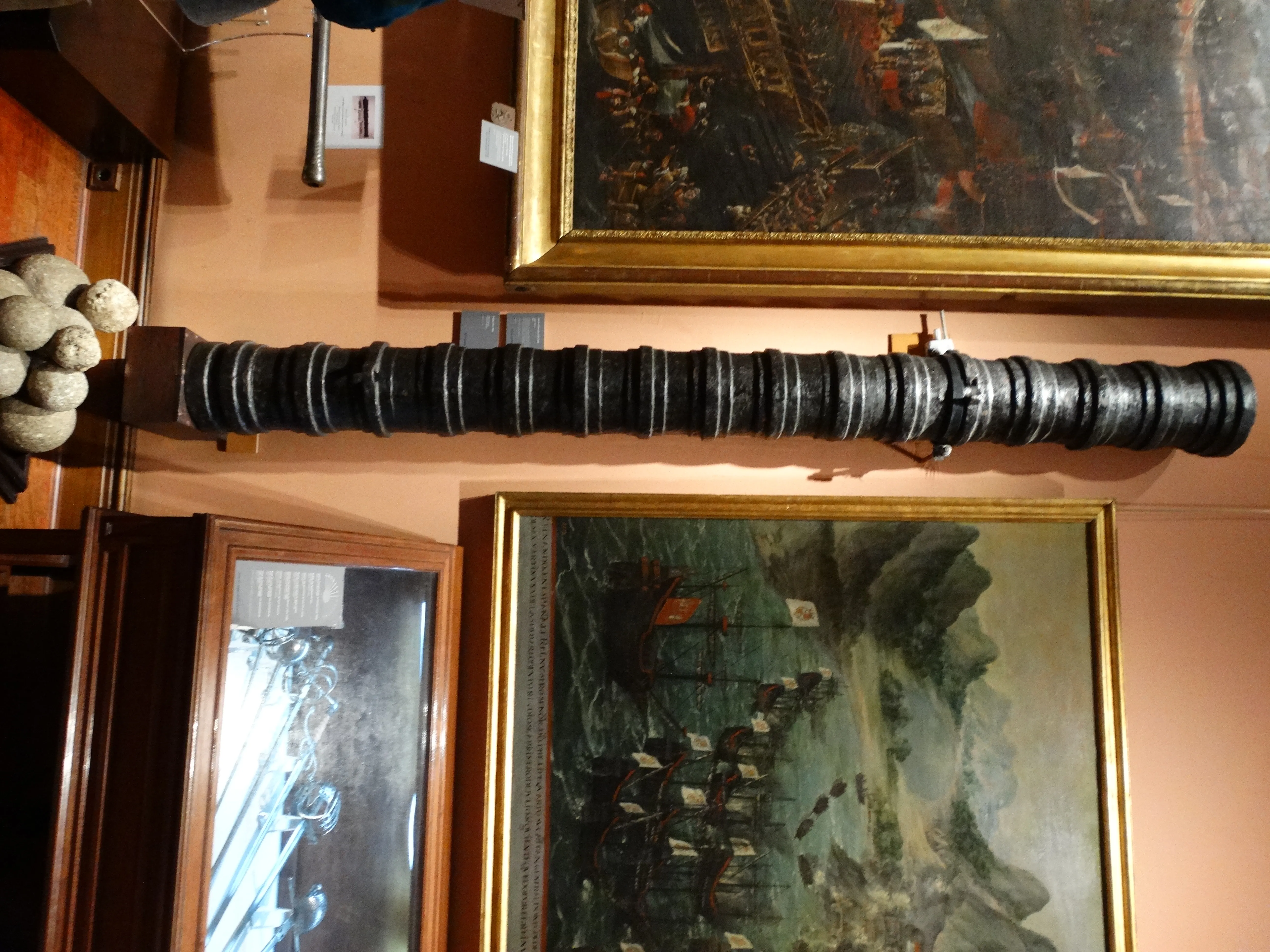 A long dark cannon on display in a museum, with paintings and artifacts in the background