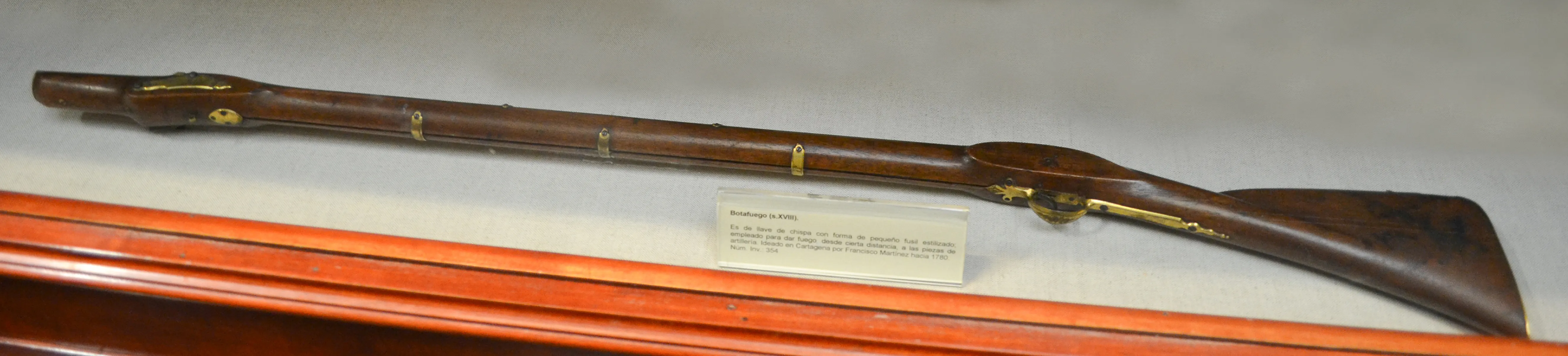 A wooden musket with brass fittings displayed in a glass case with a label