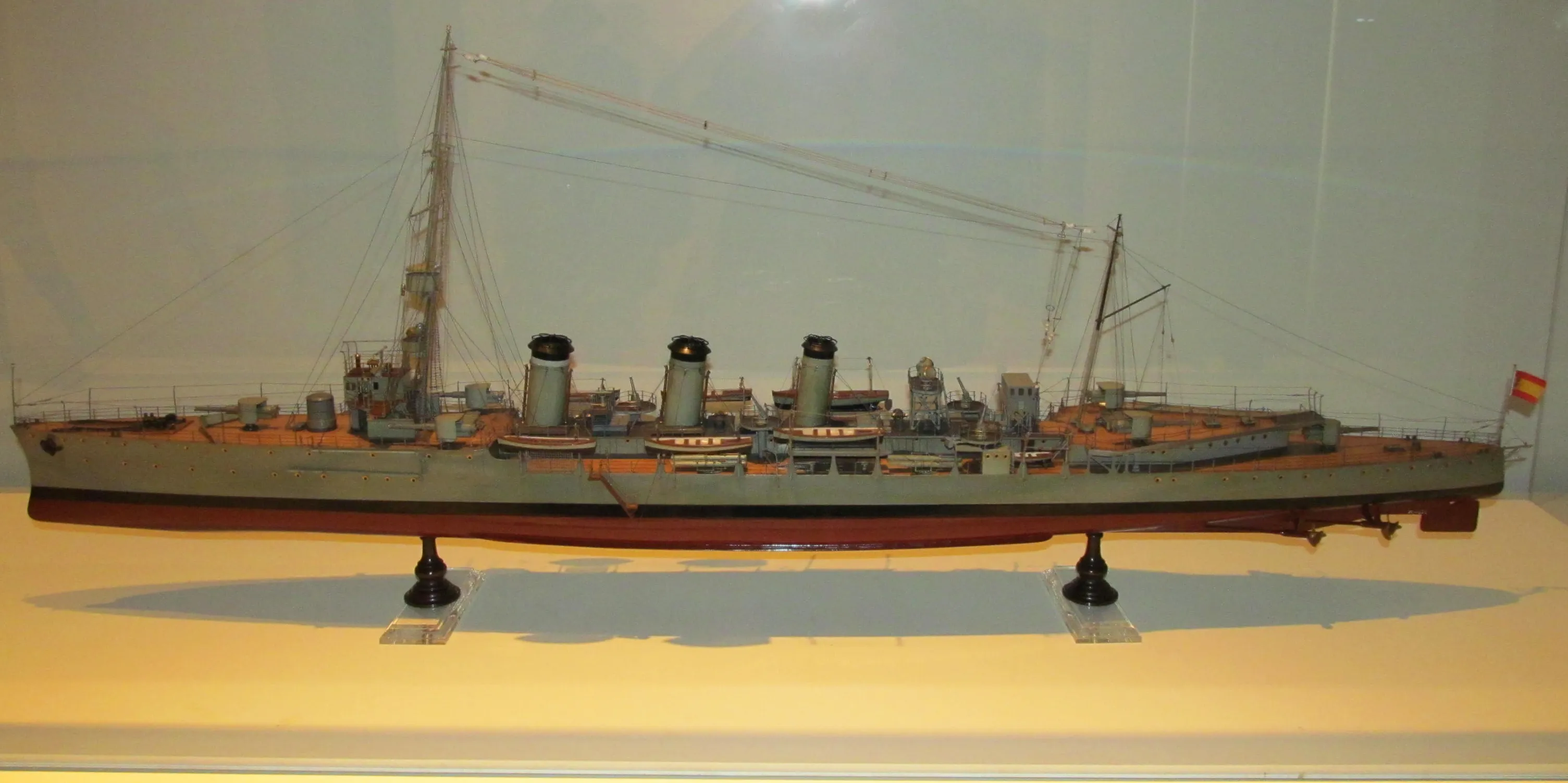 A scale model of a naval ship with multiple smokestacks and masts displayed on a stand in a museum case.