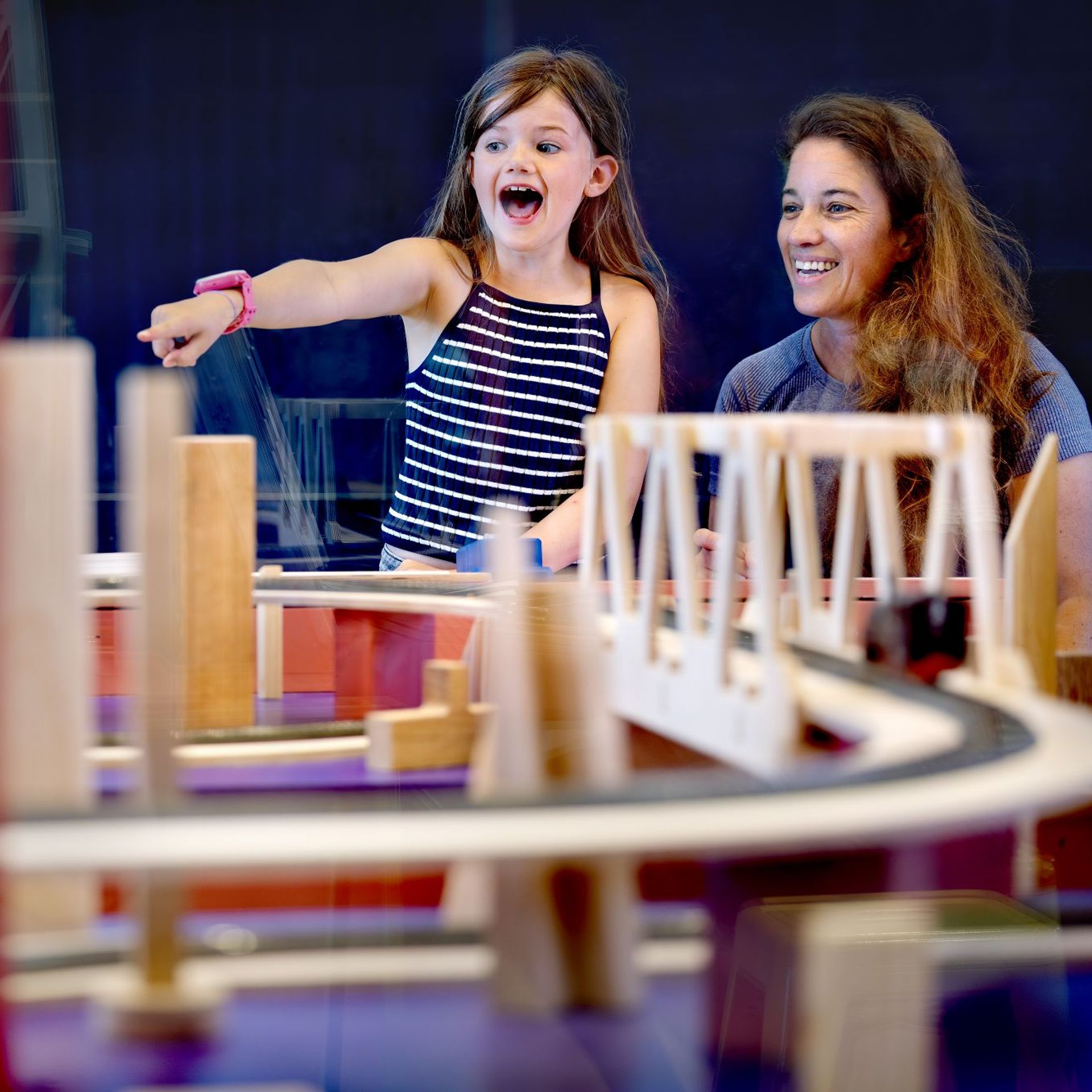 Two people interacting with a wooden bridge model exhibit featuring curved tracks and structural elements in a science museum setting