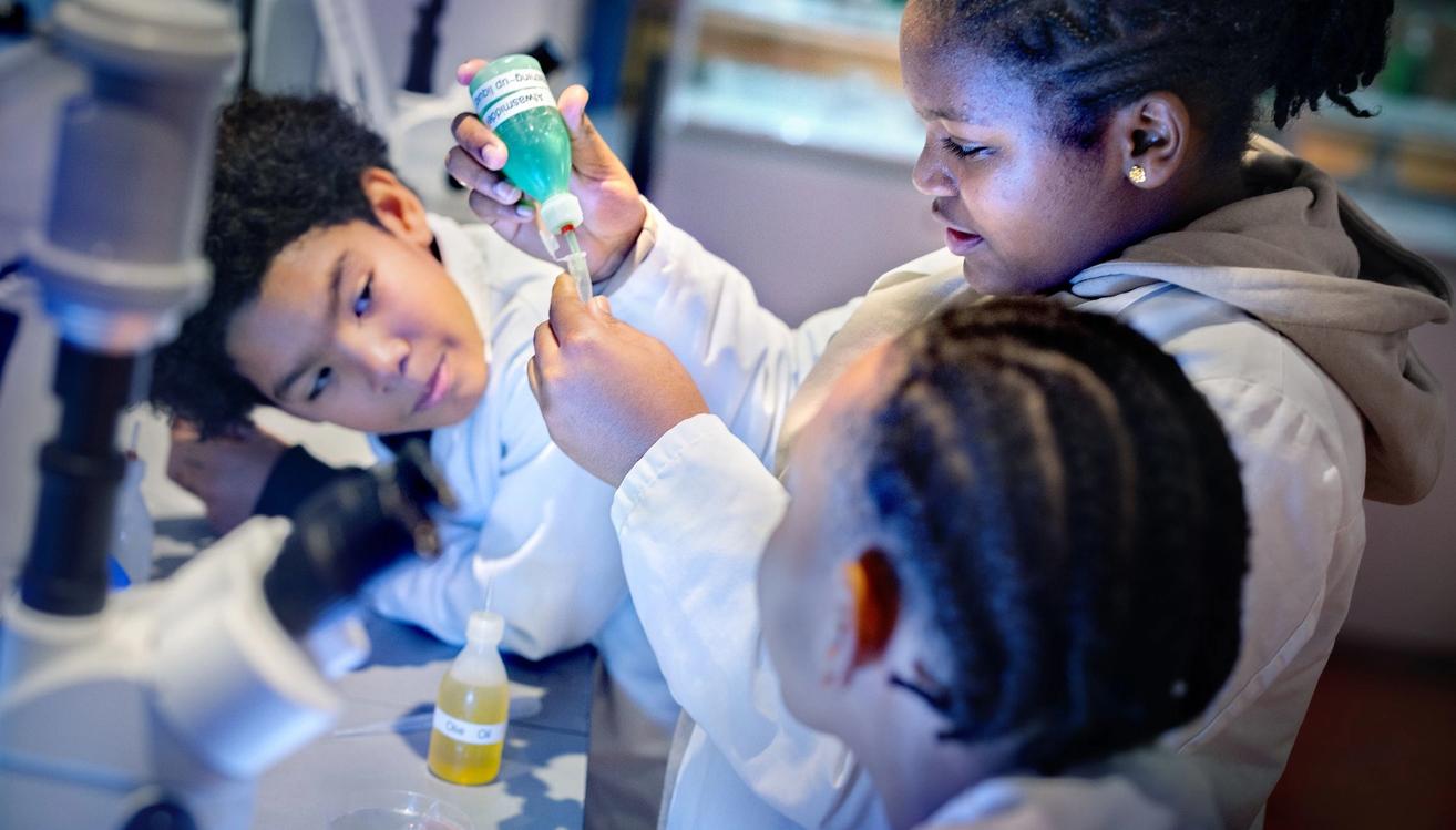 Children in white lab coats conducting science experiments with pipettes and microscopes at an interactive museum exhibit