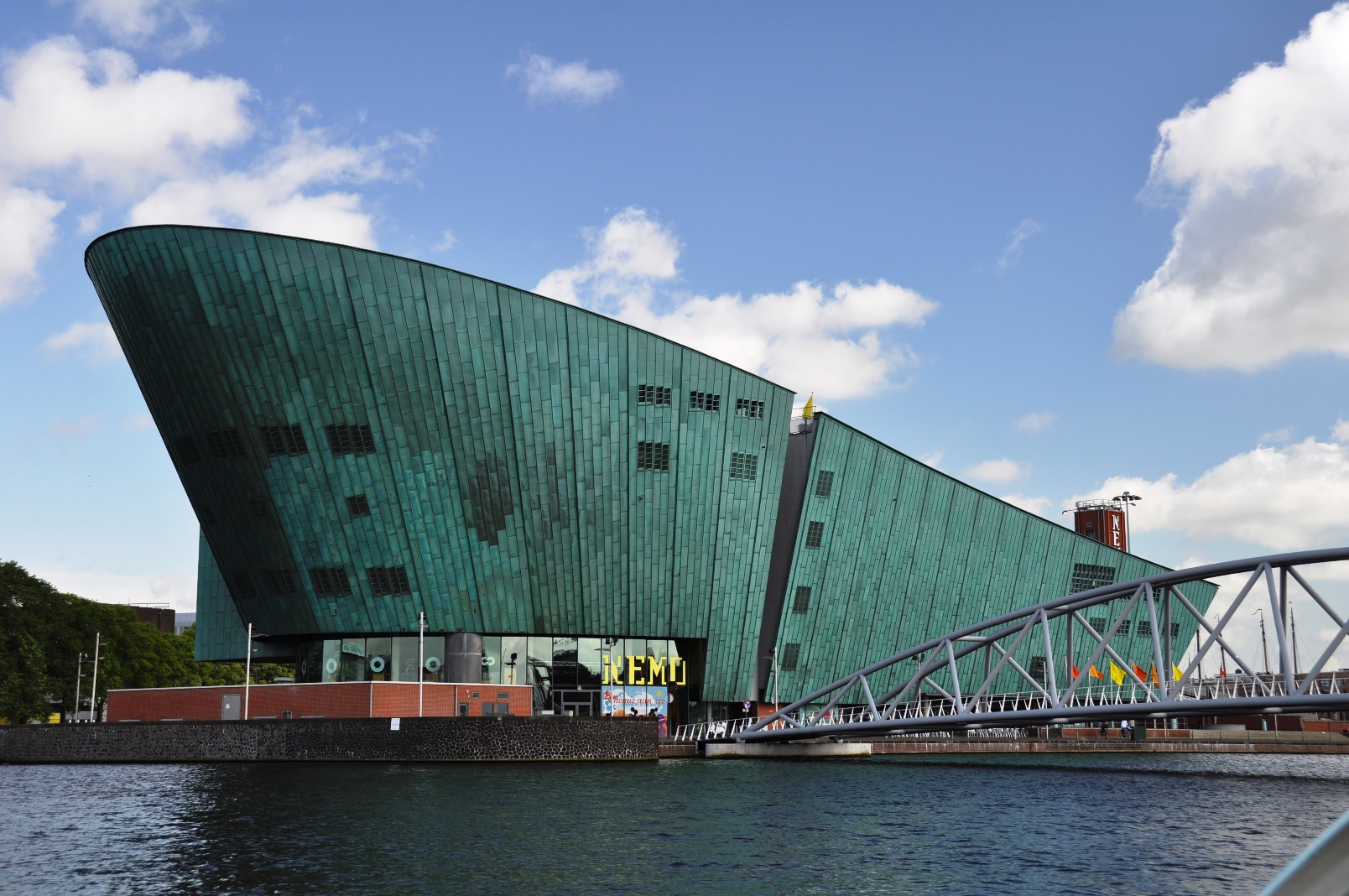 Modern gebouw met een groene, gebogen gevel en een groot bord met de tekst NEMO, gelegen naast een rivier met op de voorgrond een brug