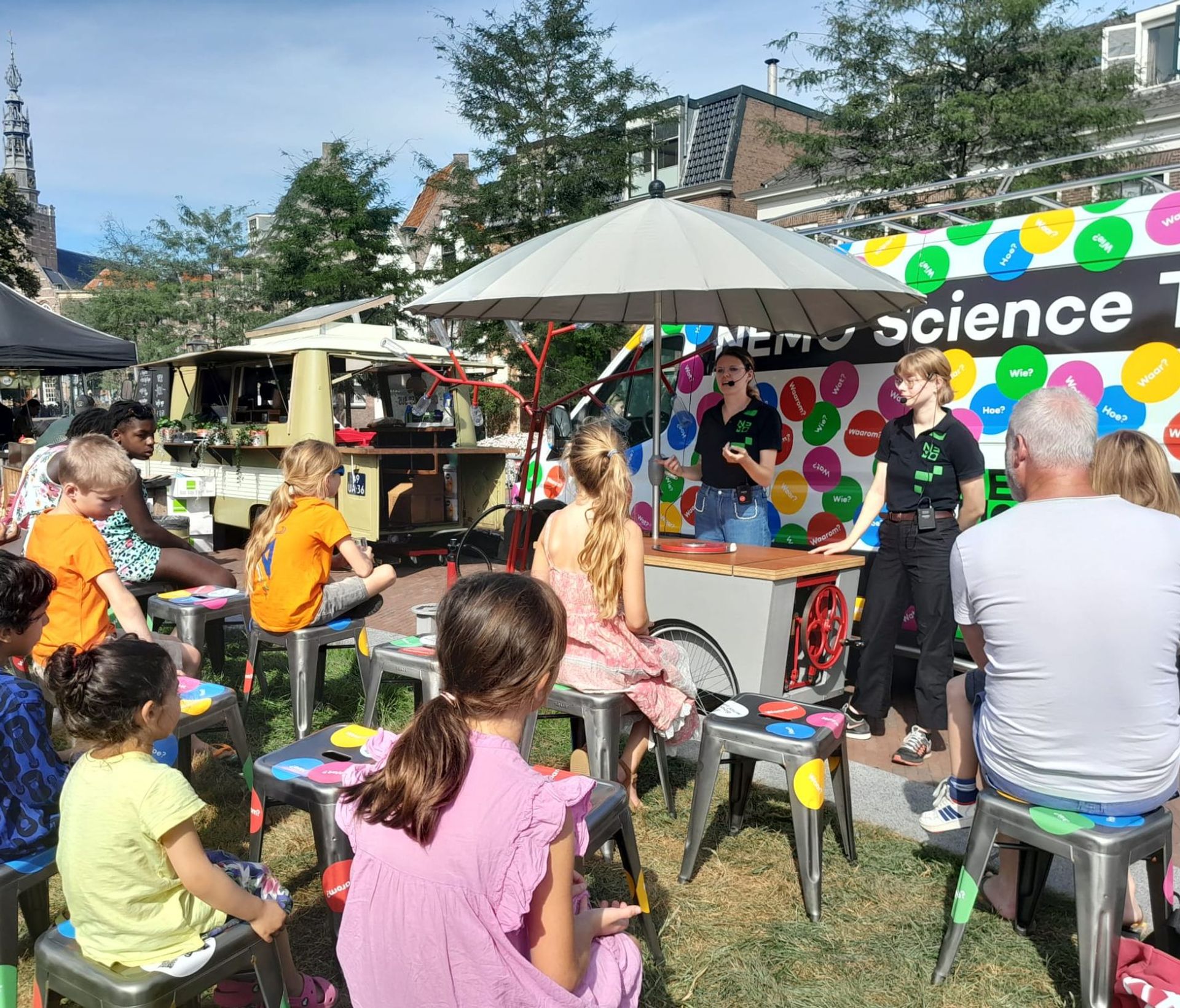 Kinderen en volwassenen zittend op kleurrijke krukjes buiten bij NEMO Science Museum, met personeel dat presenteert voor een kleurrijke achtergrond en foodtrucks op de achtergrond.