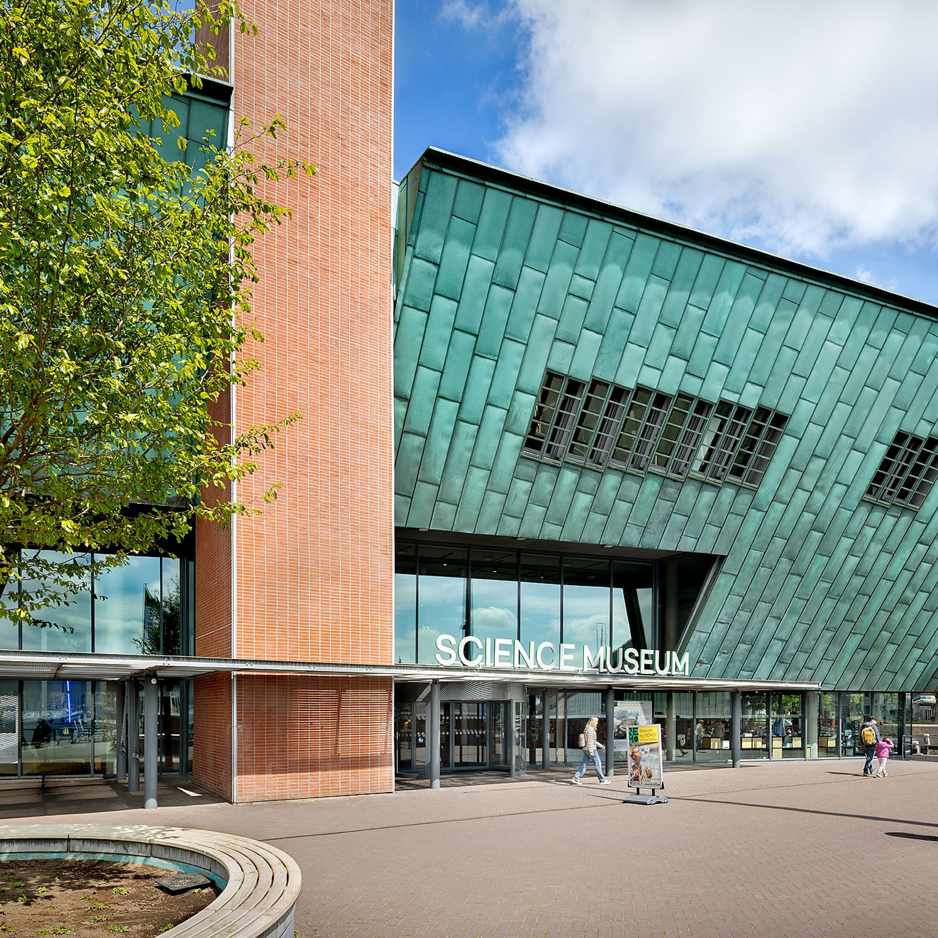 Modern gebouw met gevel van groene tegels en bakstenen zuil, ingang met 'SCIENCE MUSEUM' uithangbord, mensen lopen buiten op een zonnige dag