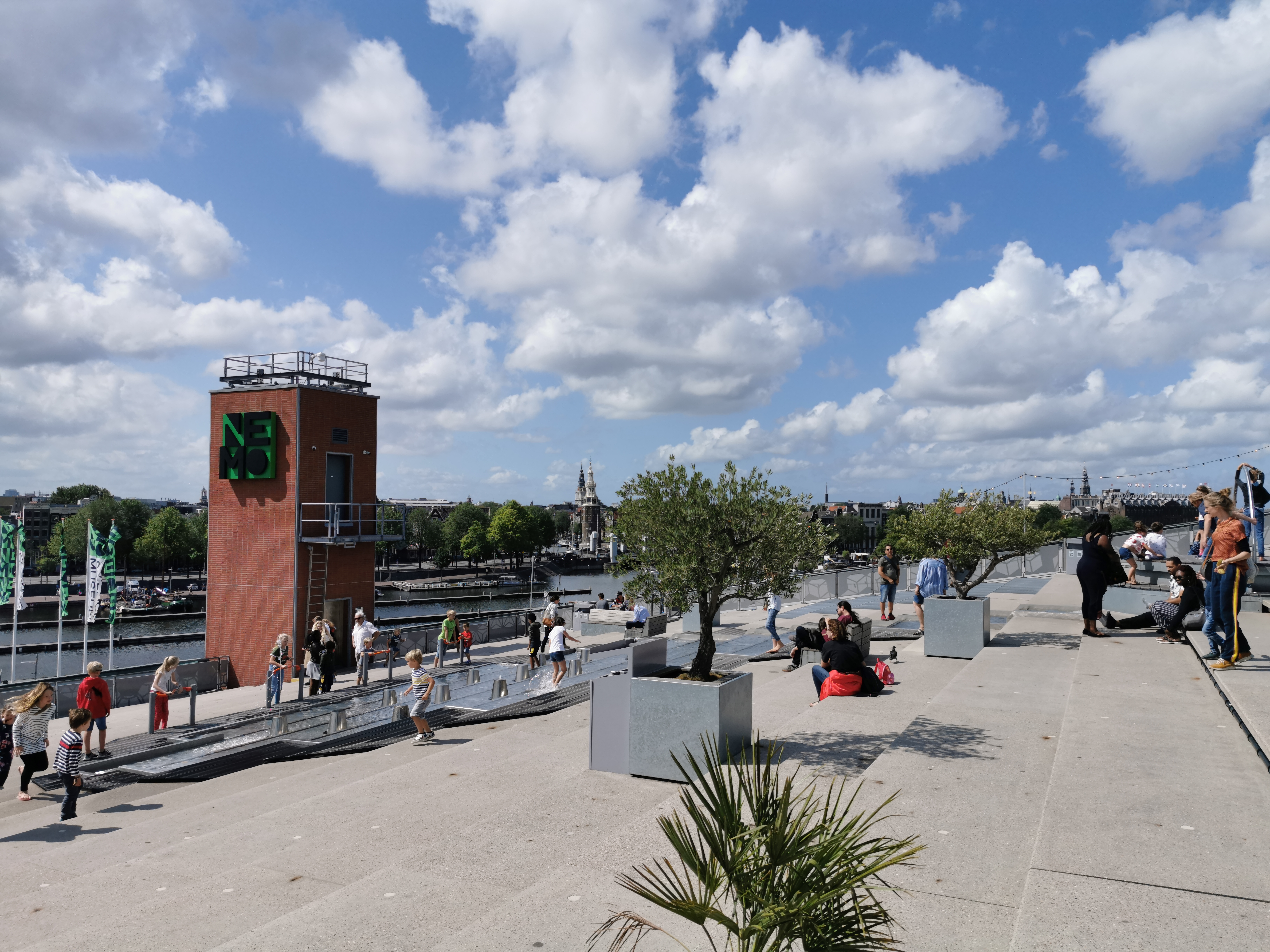 Rooftop terrace with people gathered near a red brick NEMO tower, trees in planters, and a city skyline under a partly cloudy sky