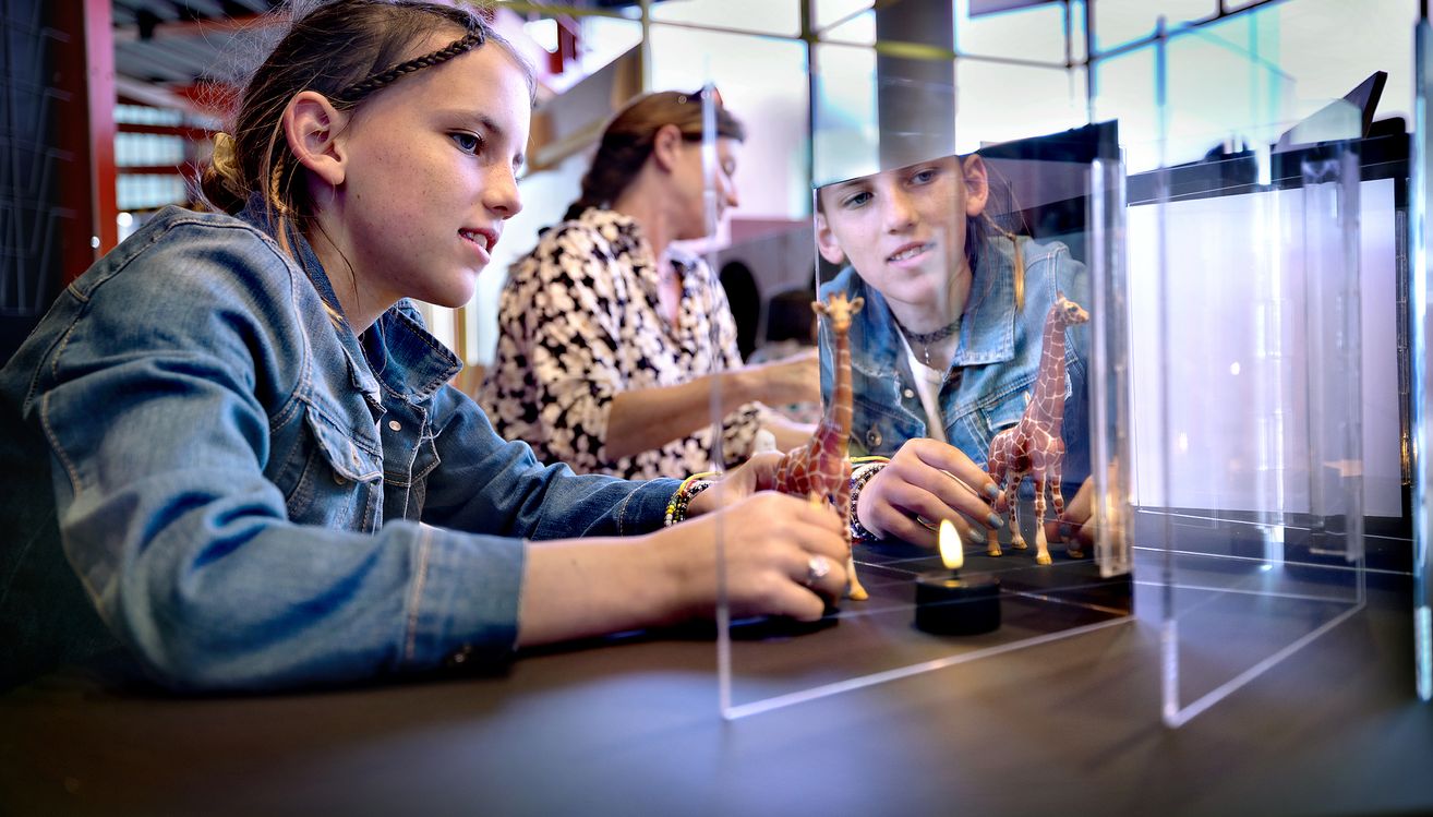 Kinderen die interageren met een wetenschappelijke tentoonstelling met girafmodellen en een vlam in een glazen omheining in het museum.
