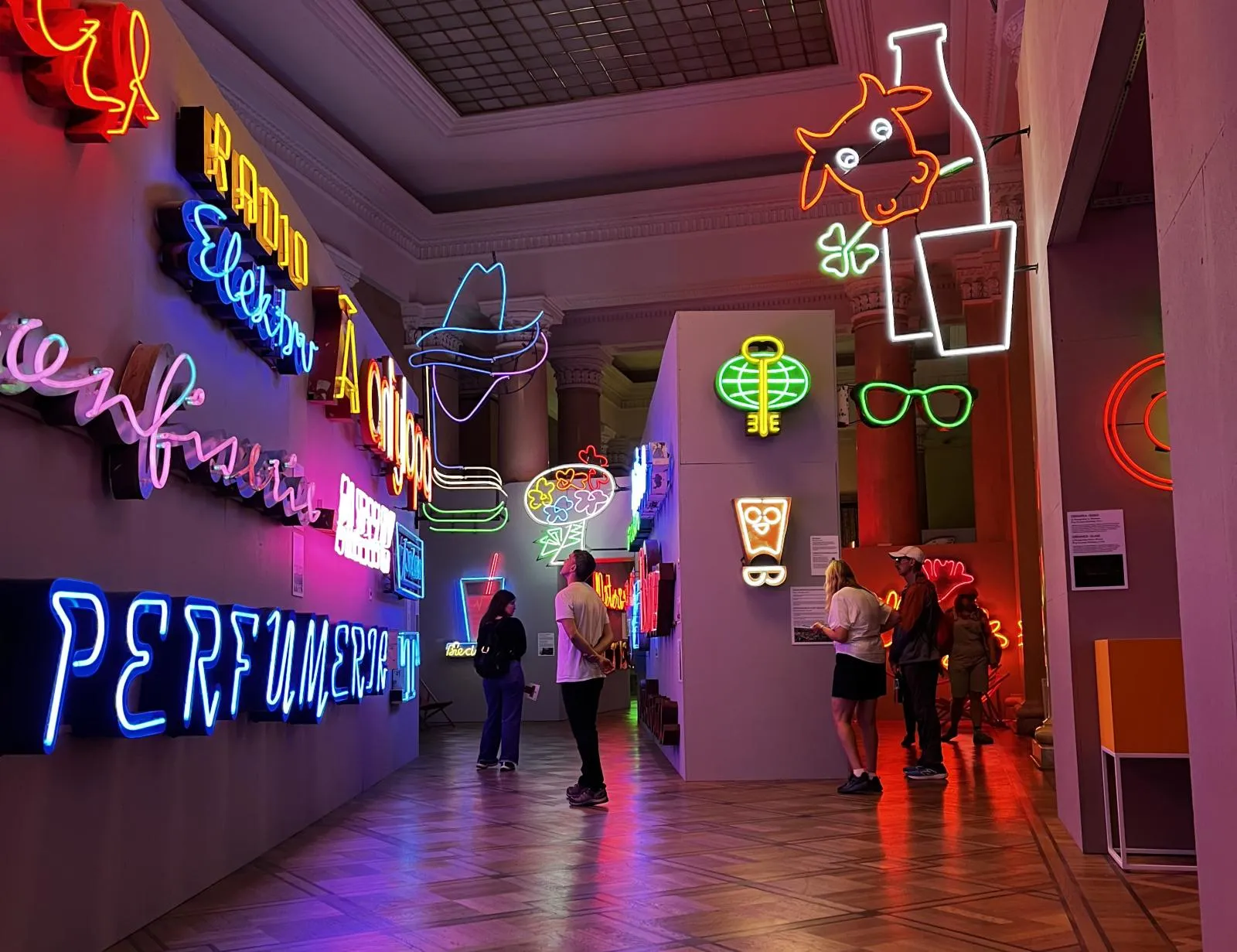 Neon signs in various colors and shapes displayed in a museum gallery with visitors walking through