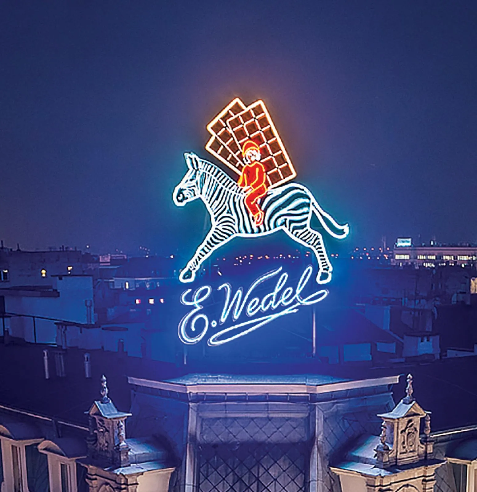 Neon sign of a zebra with a rider and chocolate bar, with 'E. Wedel' text, lit up at night
