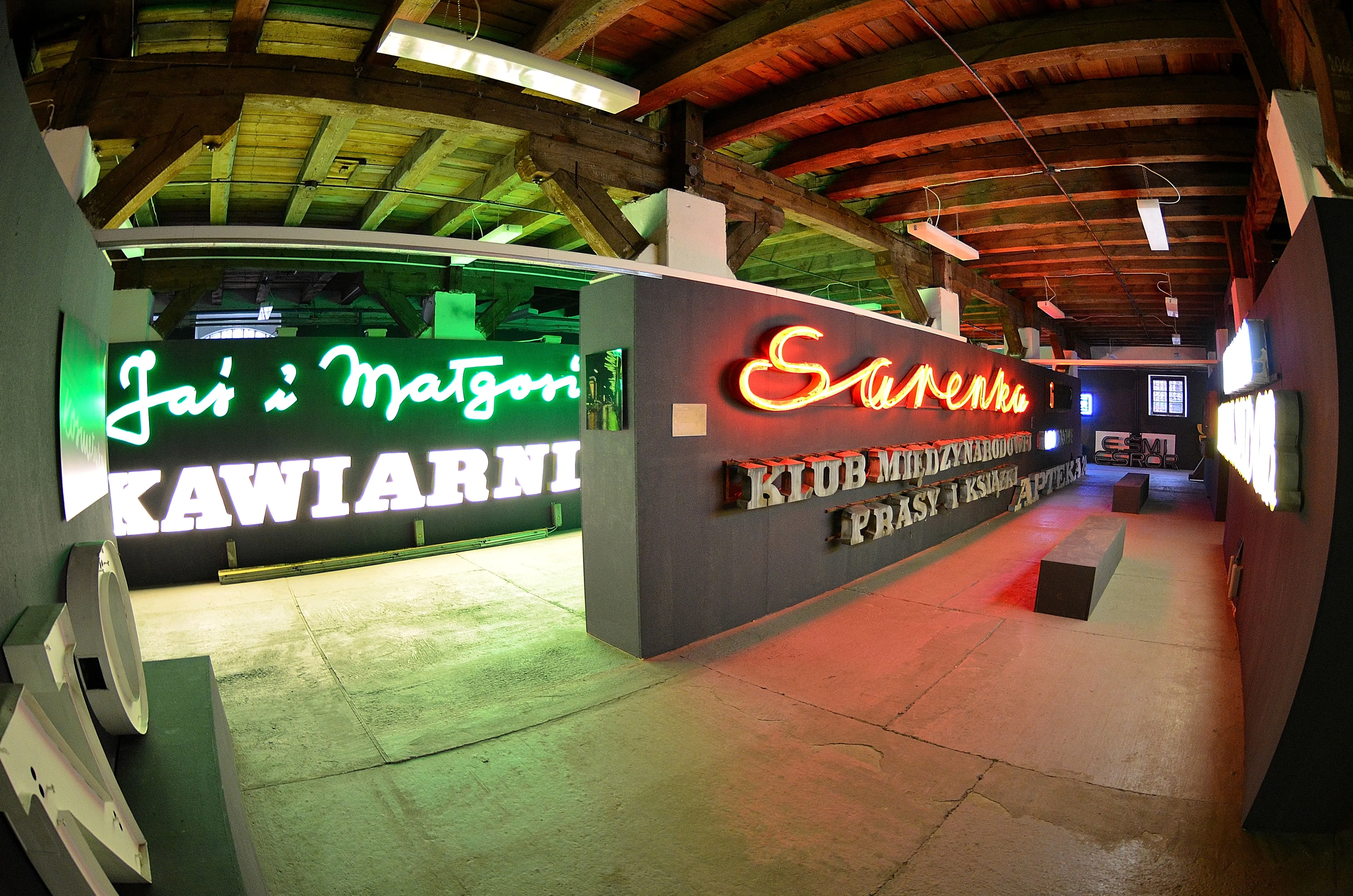 Neon signs in a museum gallery, including green and red illuminated text on dark walls under wooden beams.