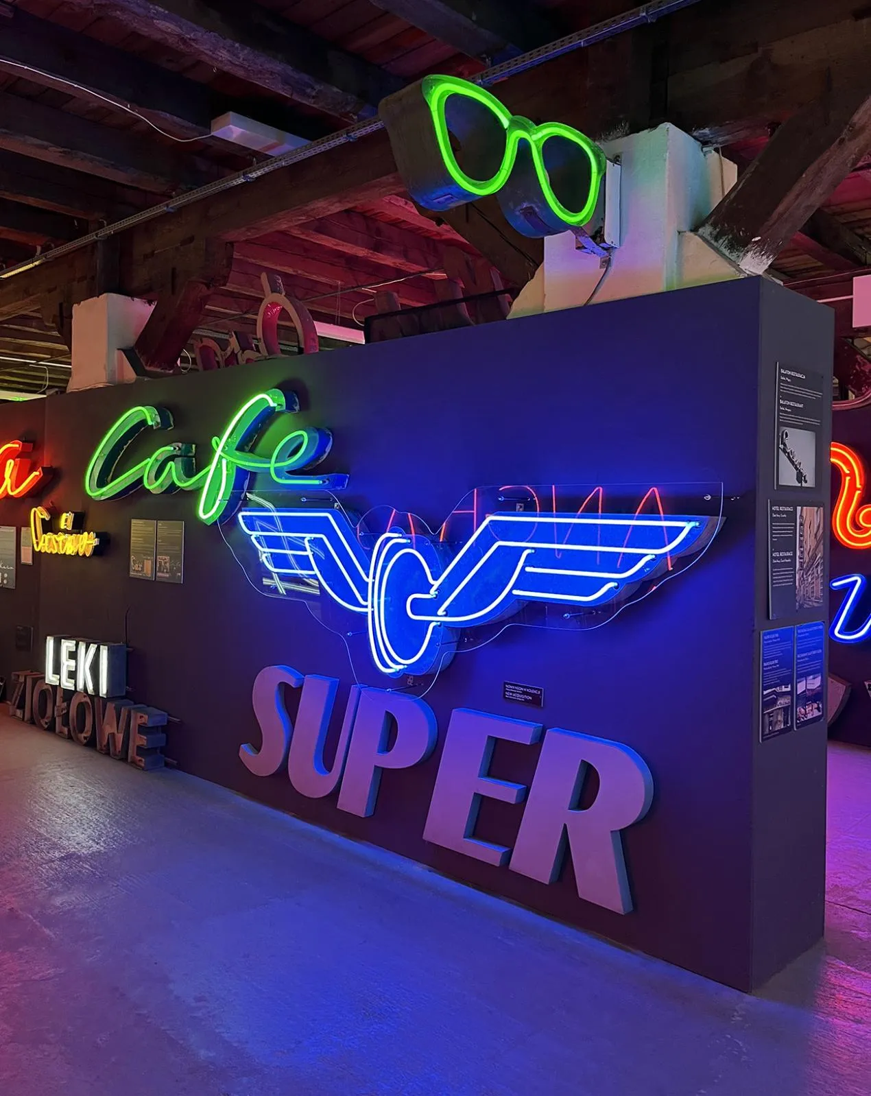 Letreros de neón que incluyen 'Cafe', un logo alado y 'SUPER' en una pared oscura dentro de un museo.