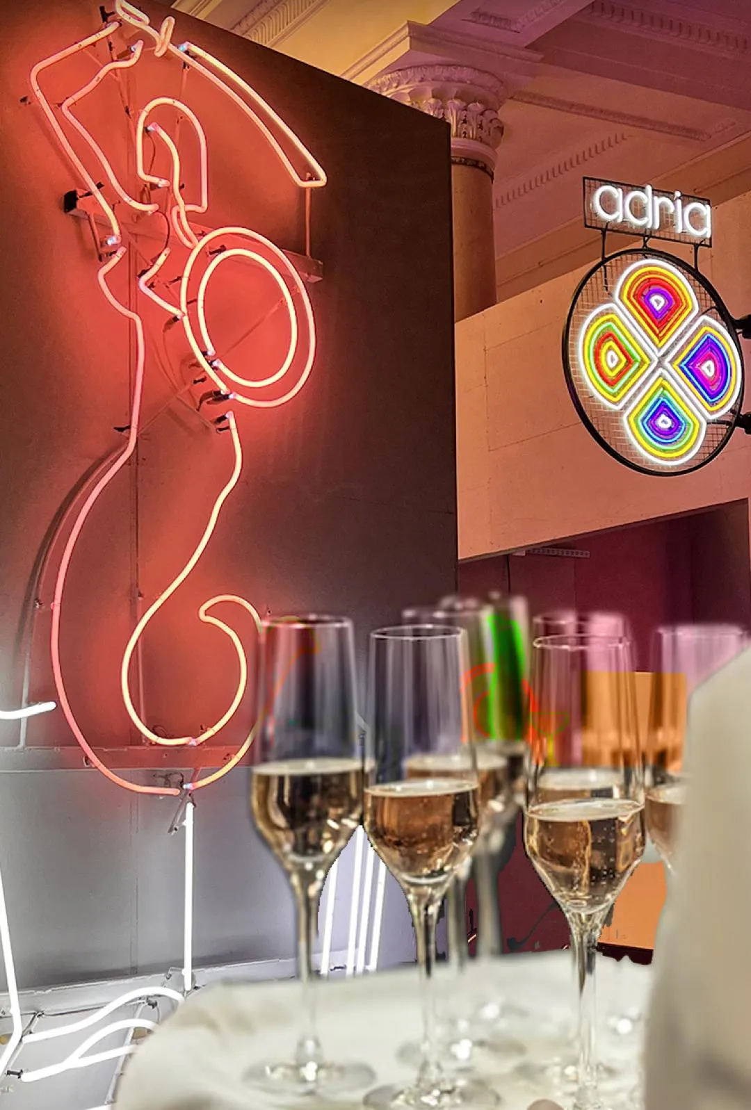 Red neon sign shaped like a figure and a circular multicolored neon sign, with champagne flutes in the foreground