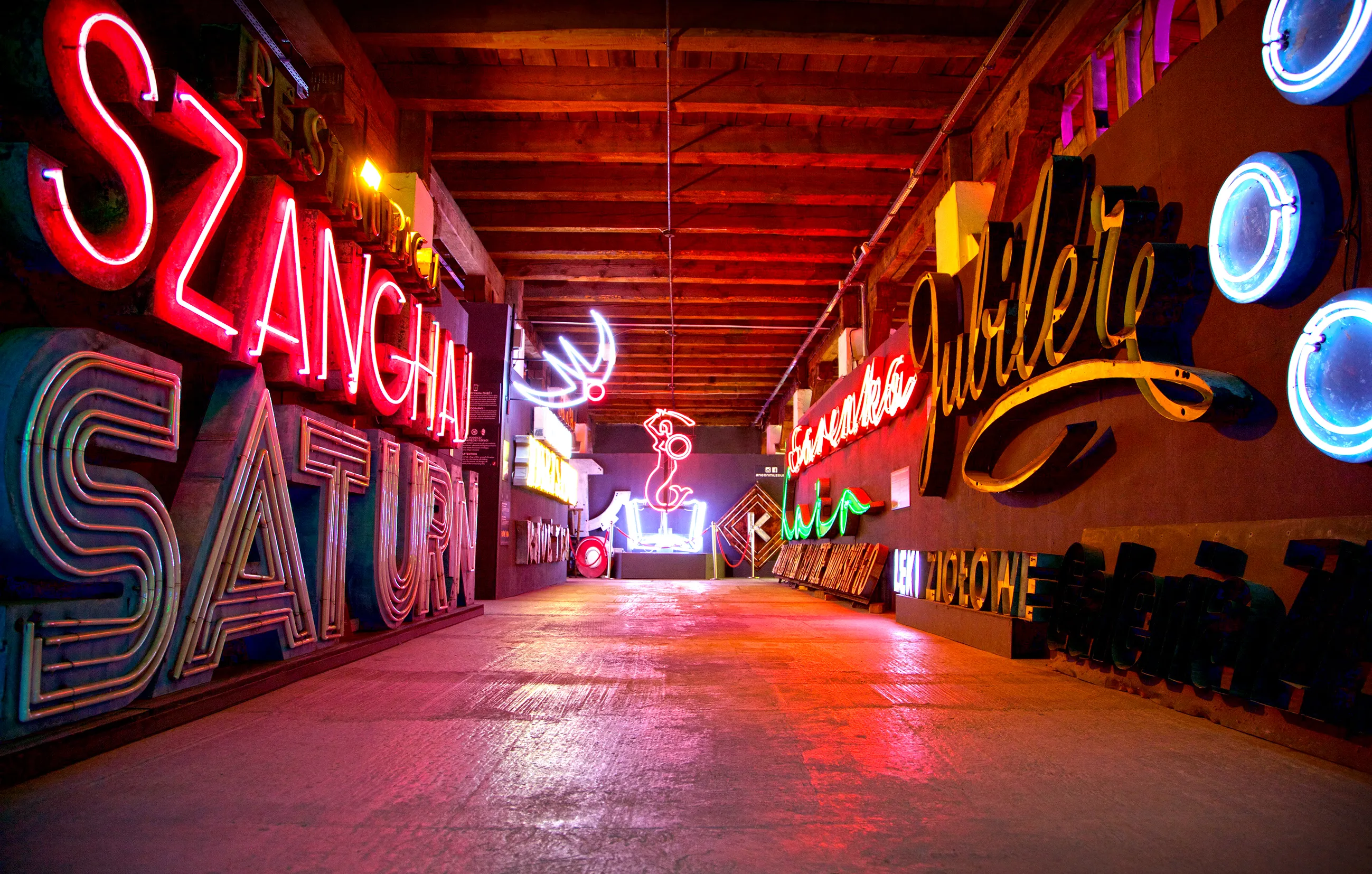 A long indoor corridor lined with glowing neon signs in various colors and styles, including red, blue, and green, with reflections on the floor.