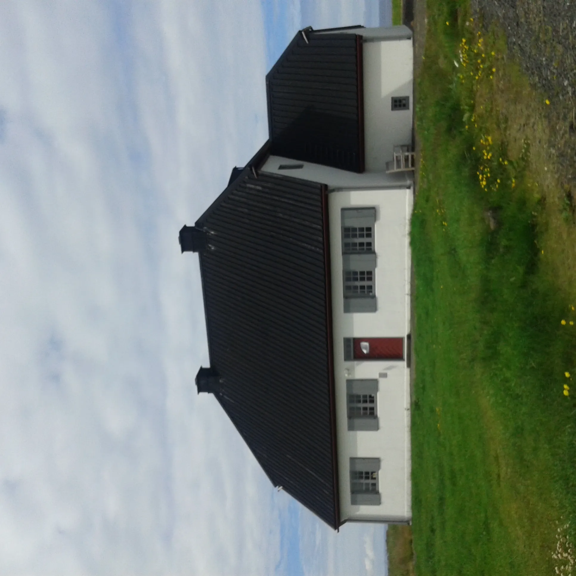 White historic stone building with dark roof and gray shutters, viewed from outside with green grass and cloudy sky