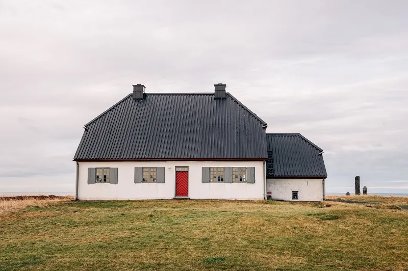 White single-story building with a dark corrugated roof and a red door, set on a grassy field under an overcast sky