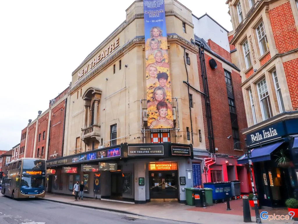 Widok z zewnątrz na New Theatre Oxford z kremową fasadą, dużym pionowym banerem z informacjami o spektaklu, oświetlonym wejściem oraz niebieskim autobusem przejeżdżającym ulicą.