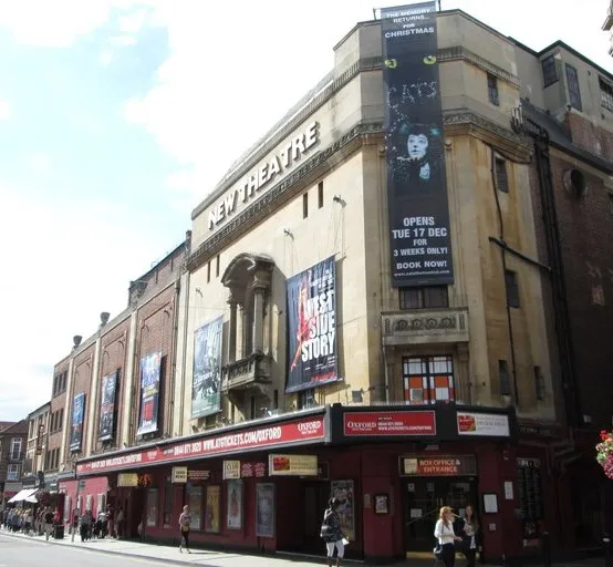 Widok z zewnątrz na budynek New Theatre Oxford przy George Street z dużymi pionowymi banerami i czerwoną fasadą.