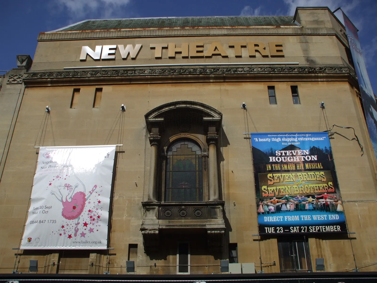 Fasada zewnętrzna New Theatre Oxford z dużym napisem NEW THEATRE na górze oraz dwoma banerami promocyjnymi wiszącymi na kamiennym budynku.