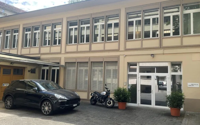 Beige gevel van een gebouw met twee verdiepingen voorzien van grote raampartijen met roedeverdeling, glazen toegangsdeur, een zwarte auto en motorfiets geparkeerd aan de voorzijde, en potplanten aan weerszijden van de ingang