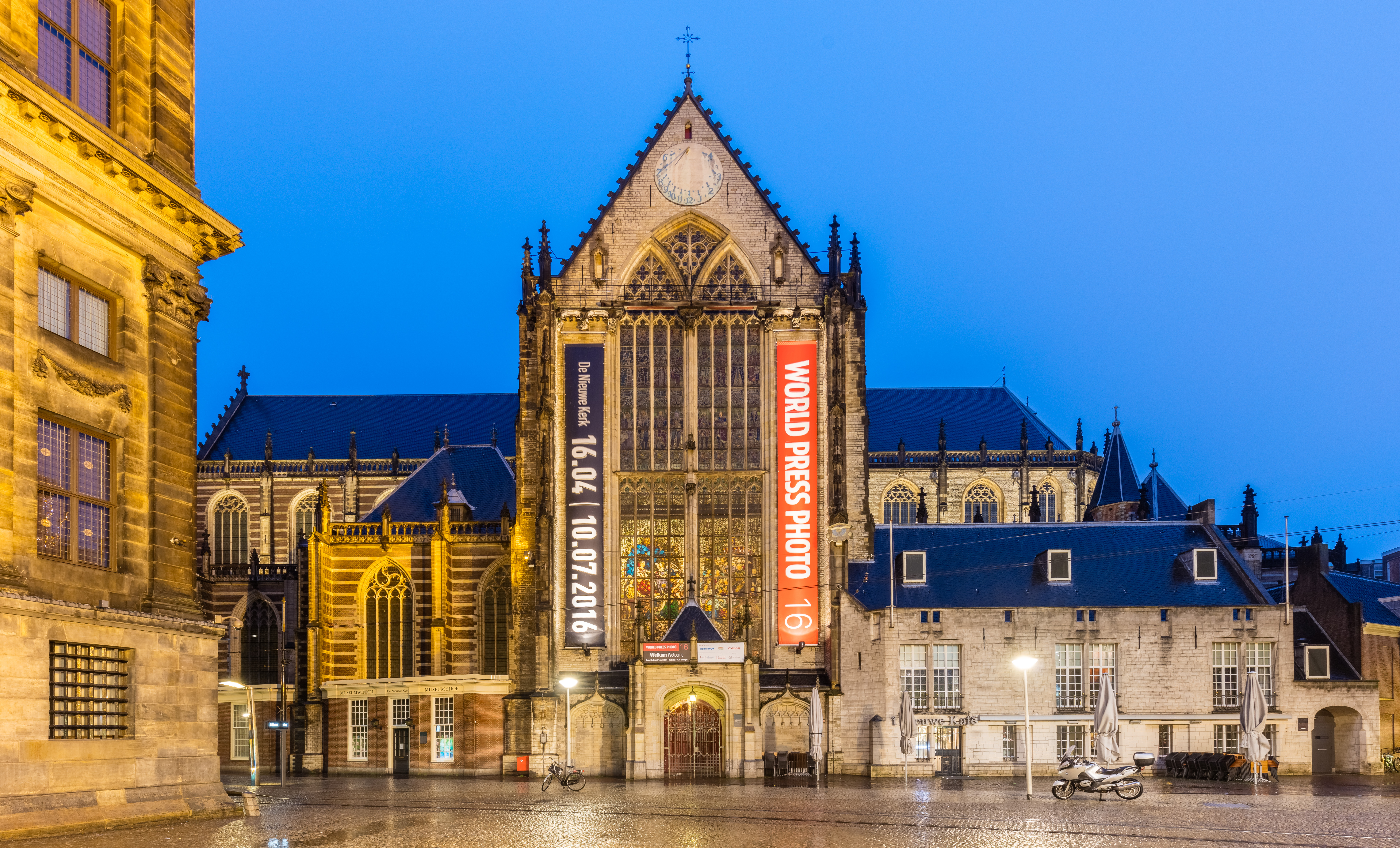 Buitenkant van de Nieuwe Kerk Amsterdam bij schemering met verlichte gotische architectuur en rode 'World Press Photo'-banners