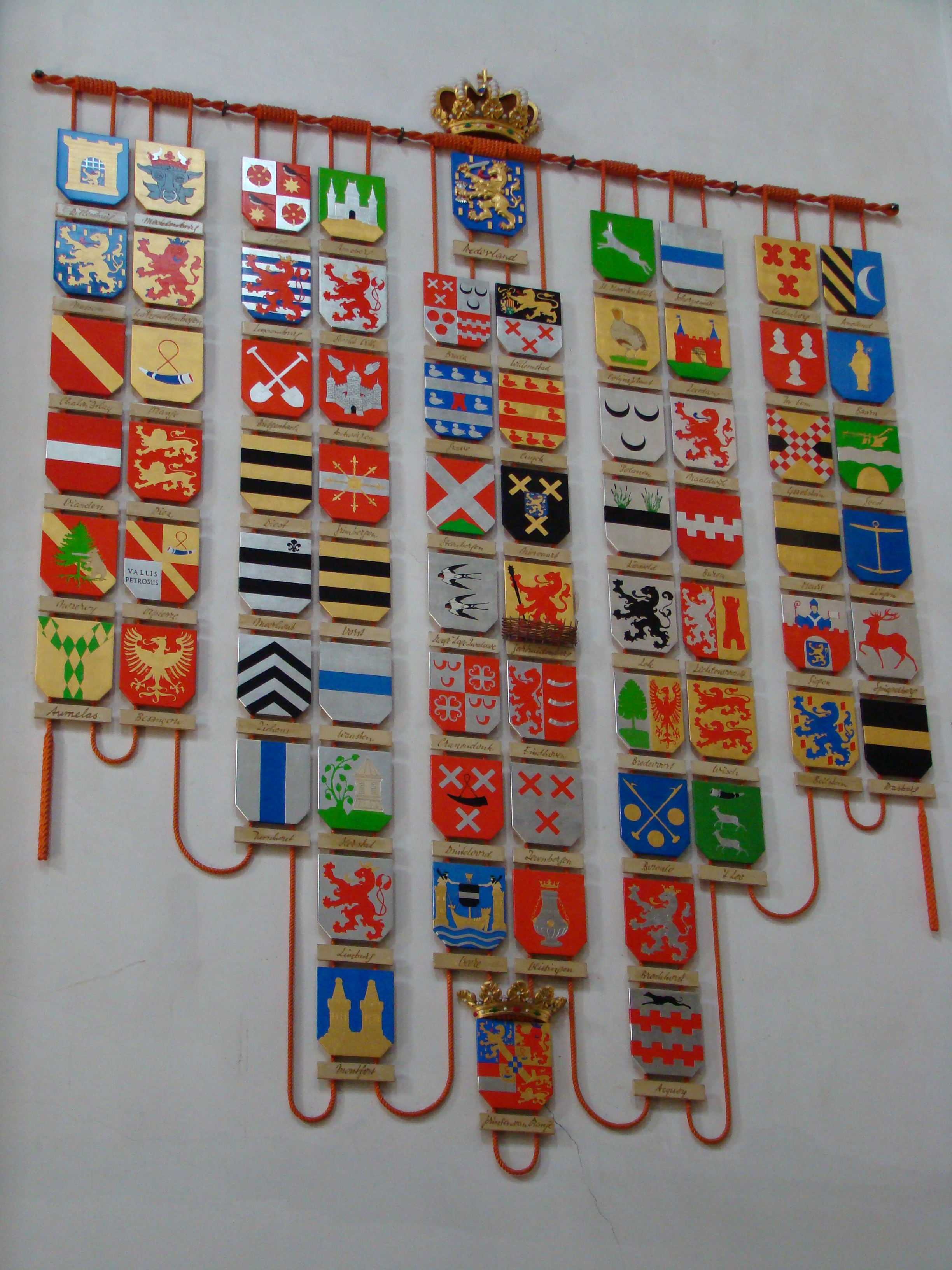 Een wanddisplay van talrijke kleurrijke heraldische schilden, gerangschikt in rijen, elk met unieke wapenschilden en symbolen, gemonteerd op een witte muur met een gouden kroon bovenaan.