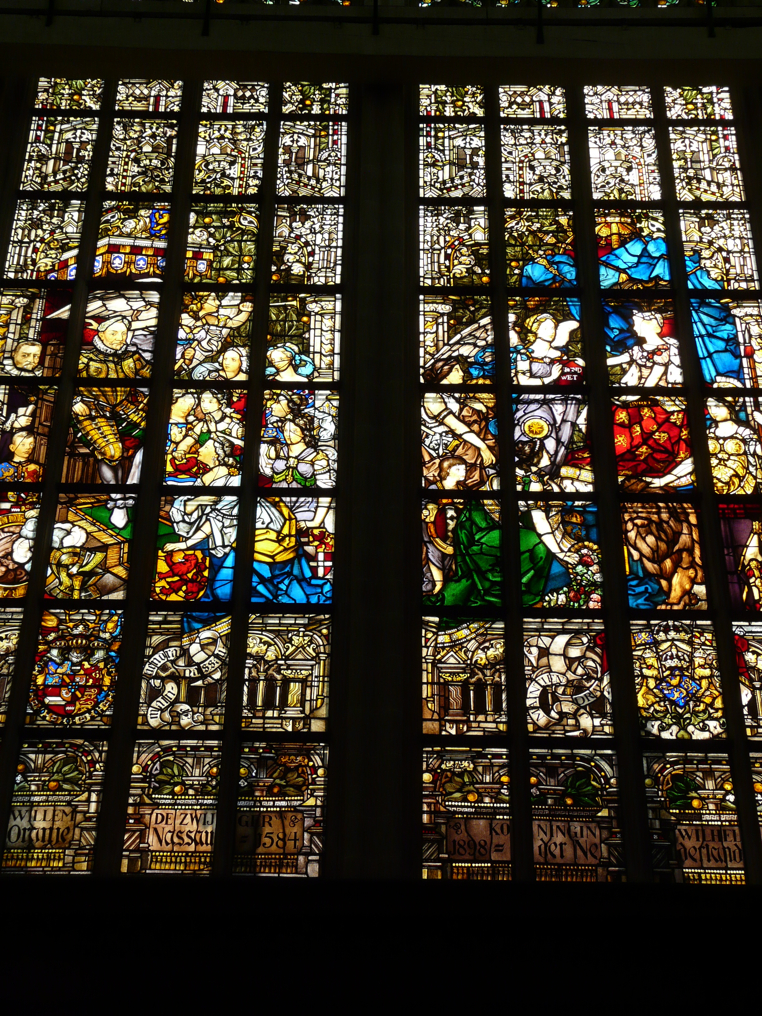 Glas-in-loodraam met ingewikkelde religieuze taferelen en figuren, verdeeld in panelen met gebrandschilderd glas, geplaatst in de Nieuwe Kerk Amsterdam.