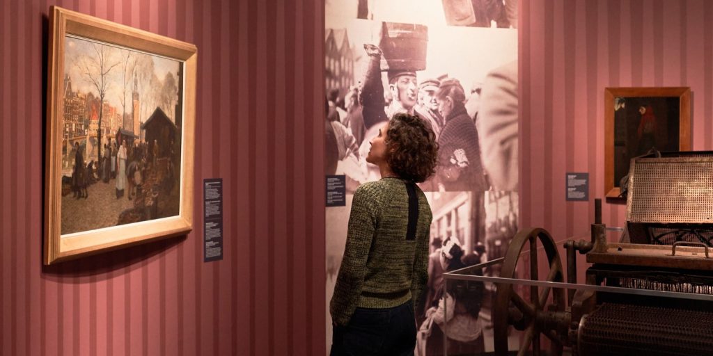 Bezoeker bekijkt historische foto's en ingelijste schilderijen in een tentoonstellingsruimte met rood gestreepte muren in De Nieuwe Kerk