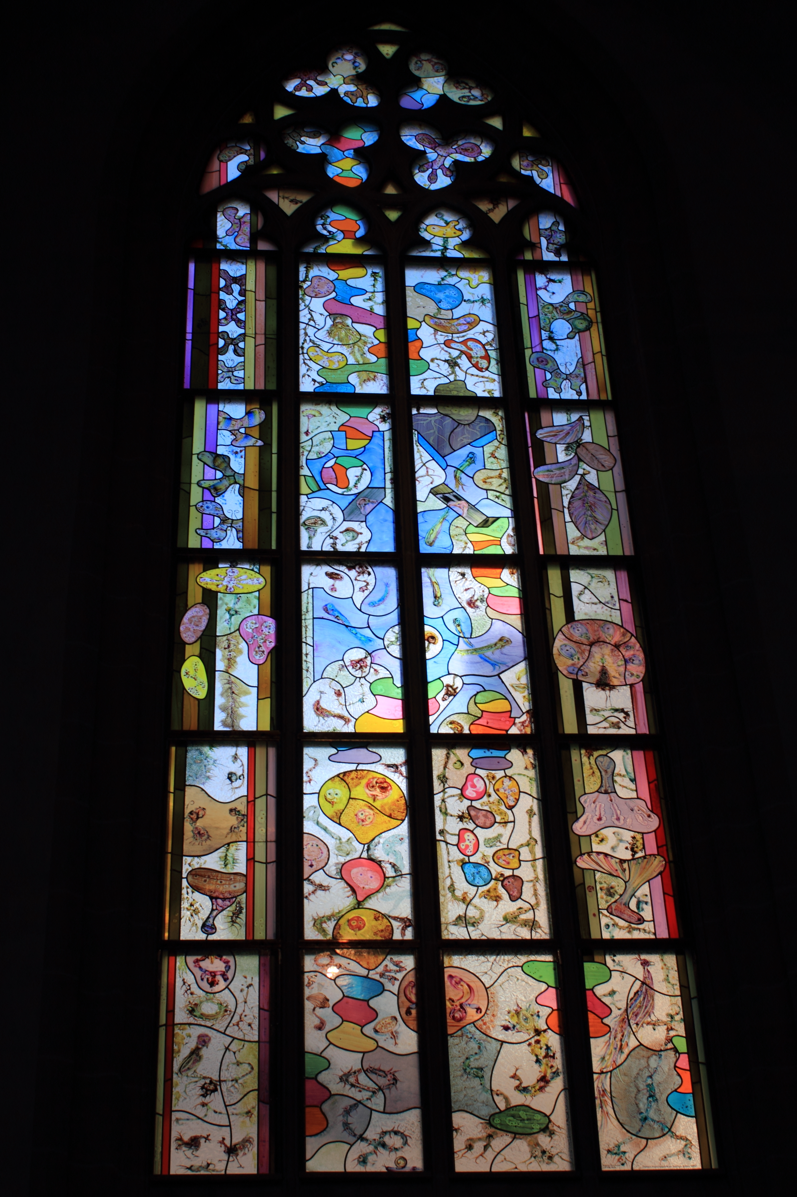 Een hoog gebrandschilderd glasraam met kleurrijke abstracte patronen en organische vormen, geplaatst in een donkere binnenruimte.