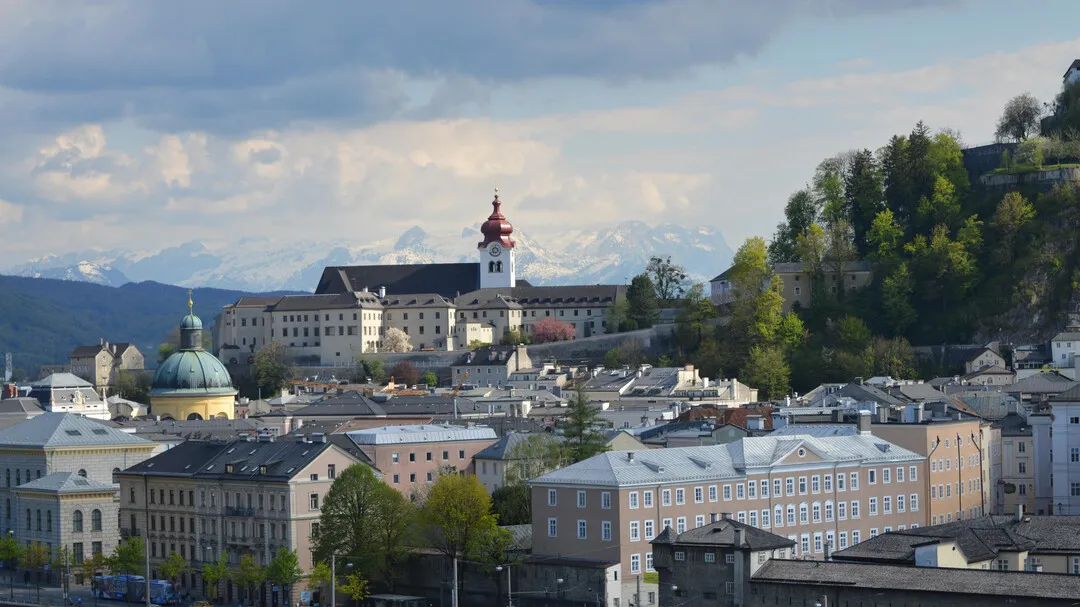 Panoramic cityscape view of Nonnberg Abbey with its red onion dome tower on a hilltop above Salzburg, surrounded by historic buildings and mountains under a cloudy sky
