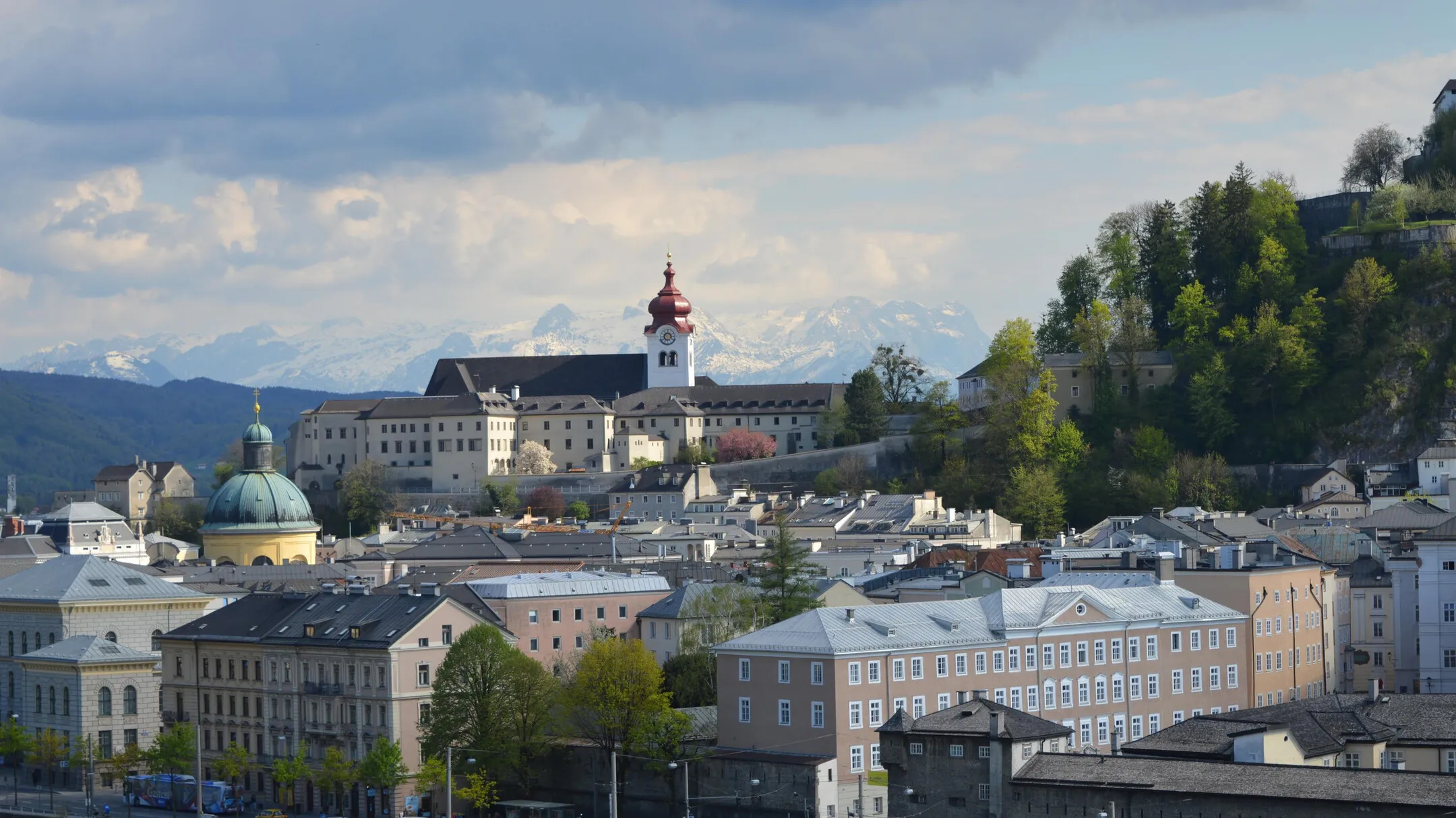 Wide cityscape view of Salzburg showing Nonnberg Abbey with its red onion dome on a hillside, surrounded by historic buildings, a green-domed church, and mountains in the background under a cloudy sky.
