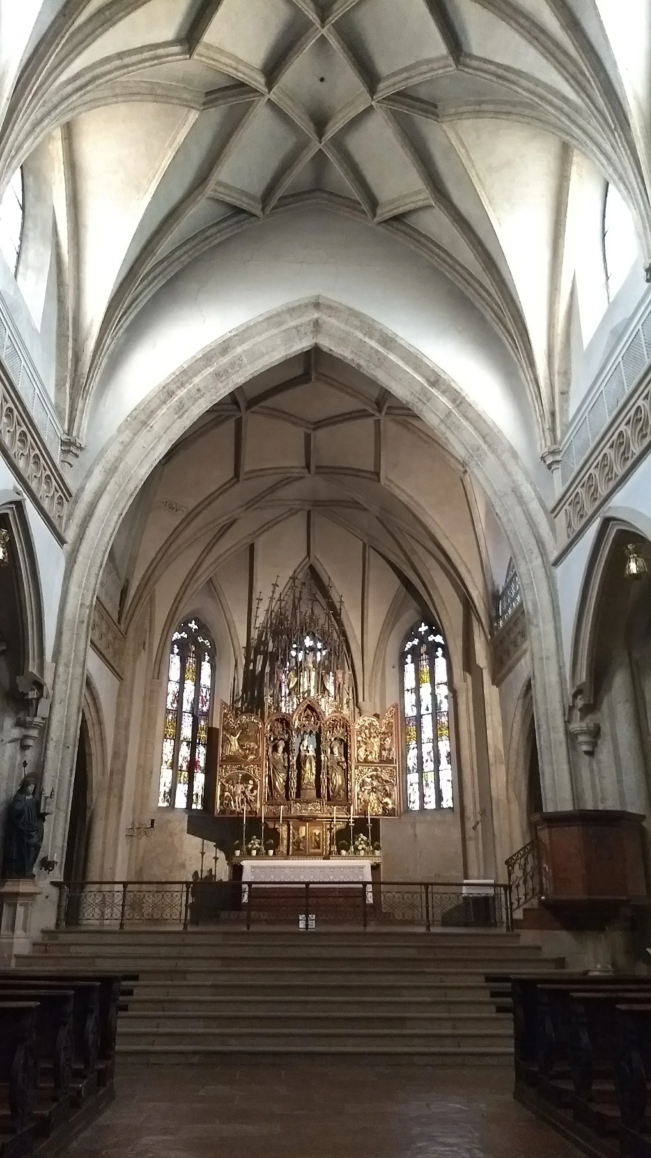Interior view of a church showing a high vaulted ceiling and a wooden Gothic altar at the far end between stained glass windows.