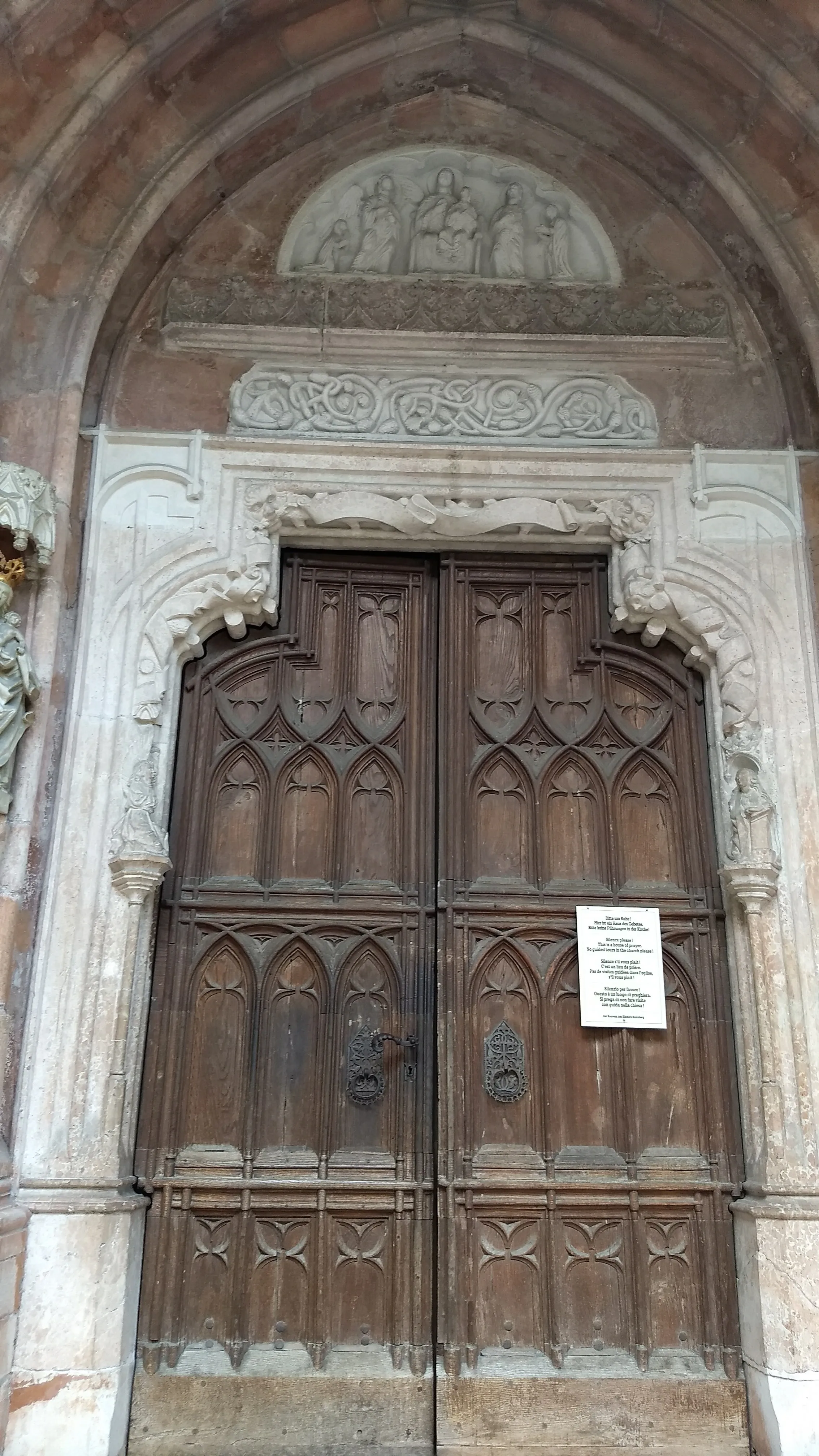 Ornate Gothic wooden double door set within a stone archway, topped with a carved relief sculpture and decorative frieze at Nonnberg Abbey