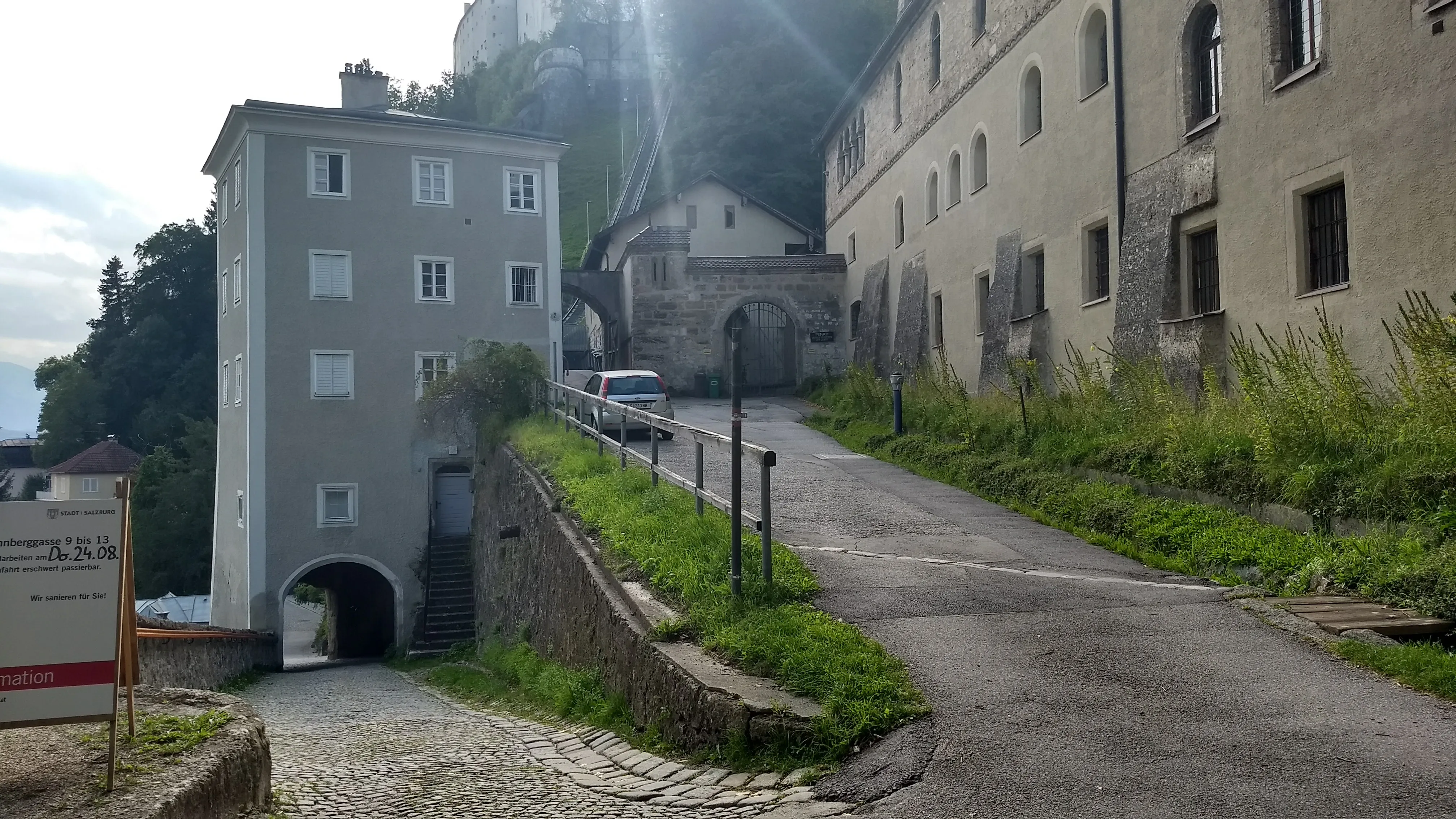 A paved road leads uphill toward a stone arched gateway flanked by historic buildings, with a tall gray multi-story building on the left and a larger stone structure on the right at Nonnberg Abbey.