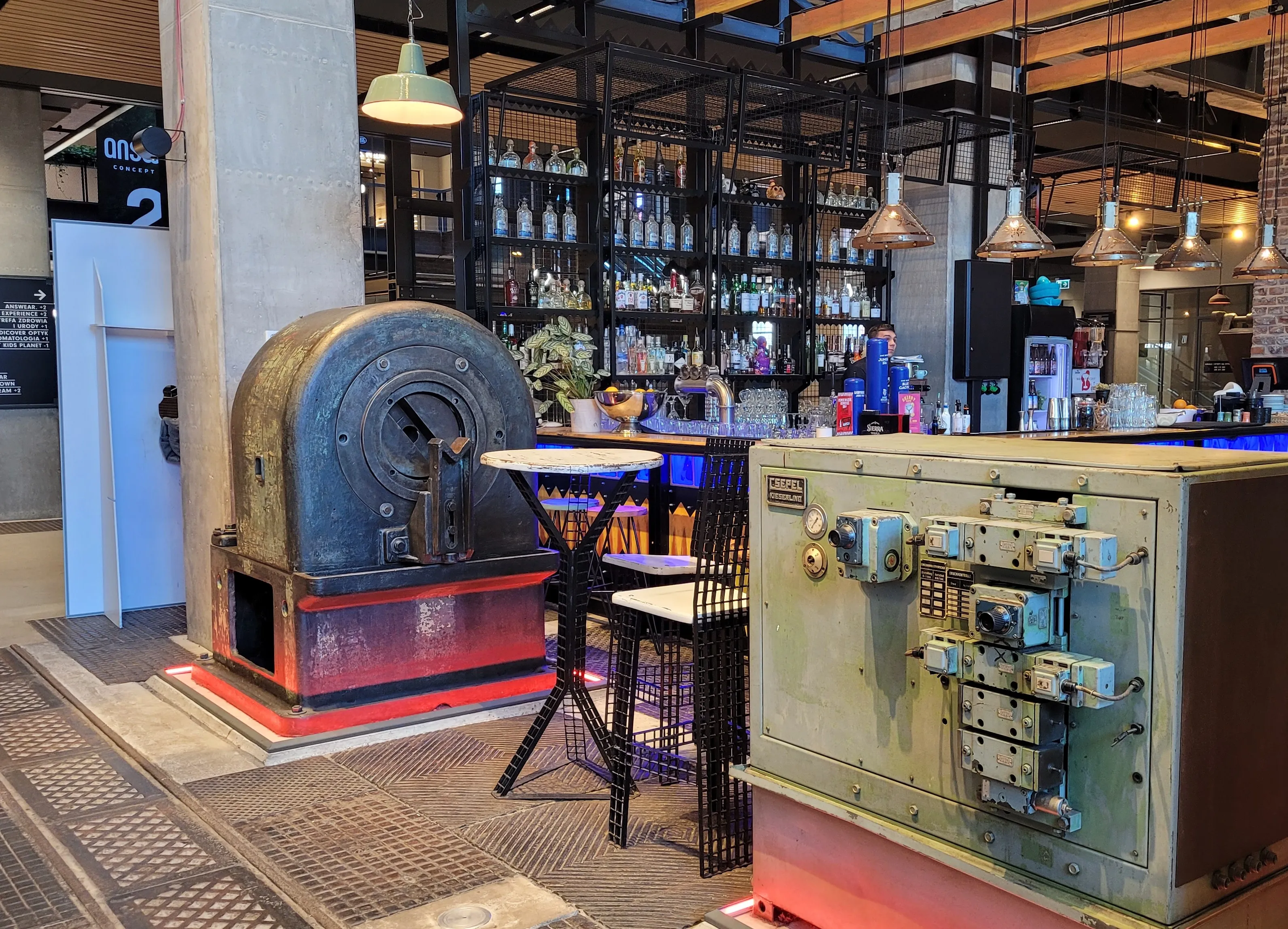 Gran máquina industrial sobre una plataforma roja junto a un panel de control vintage en un entorno de museo con estantes de botellas al fondo