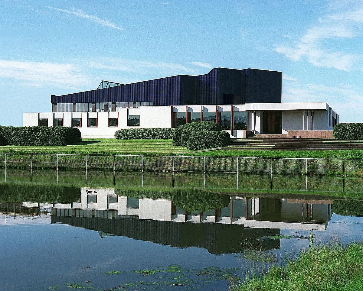 White building with dark blue upper facade reflected in a calm pond, surrounded by green hedges and lawn under a blue sky