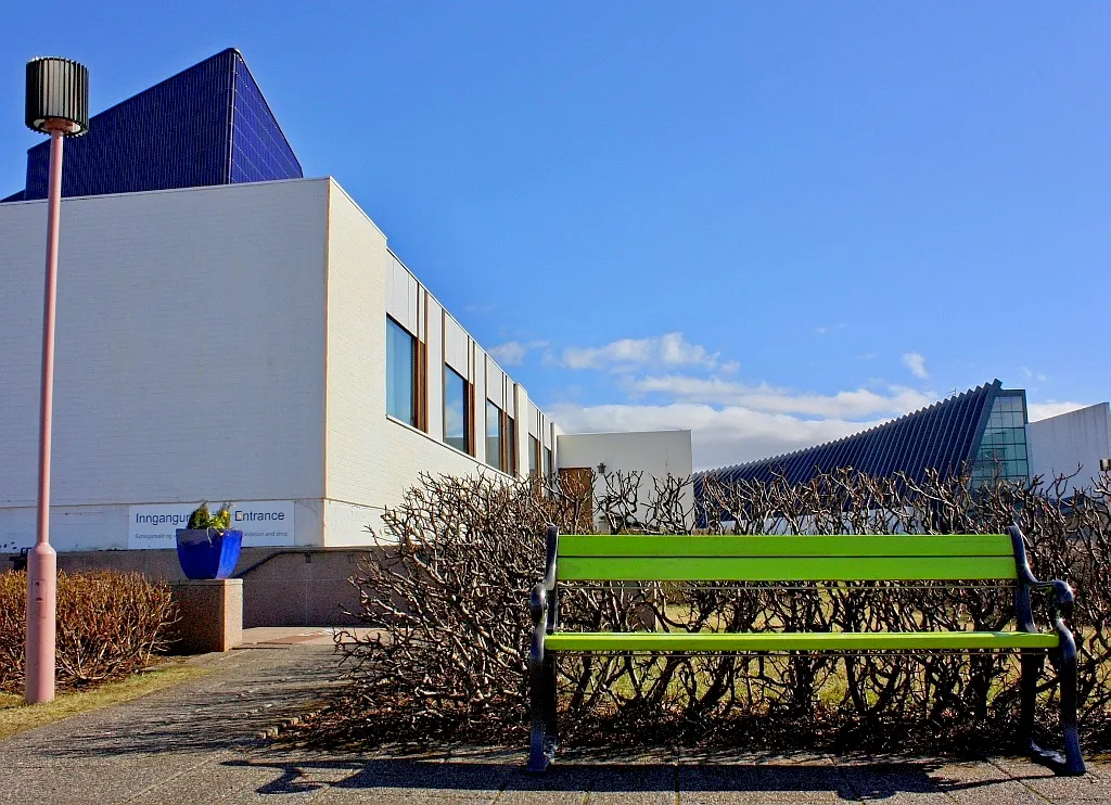 White building with a distinctive blue pyramid-shaped roof and a green bench in front of bare bushes under a clear blue sky