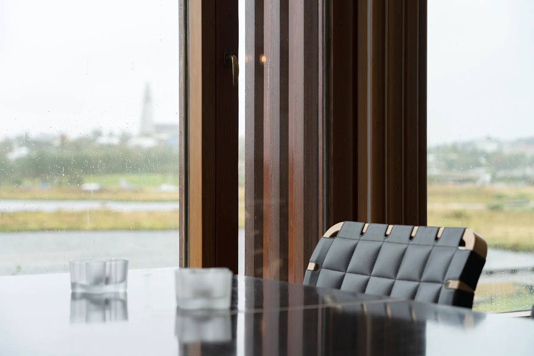 Interior of the Nordic House showing a table with glasses, a woven leather chair, and large wooden-framed windows overlooking Reykjavík with a church spire in the distance