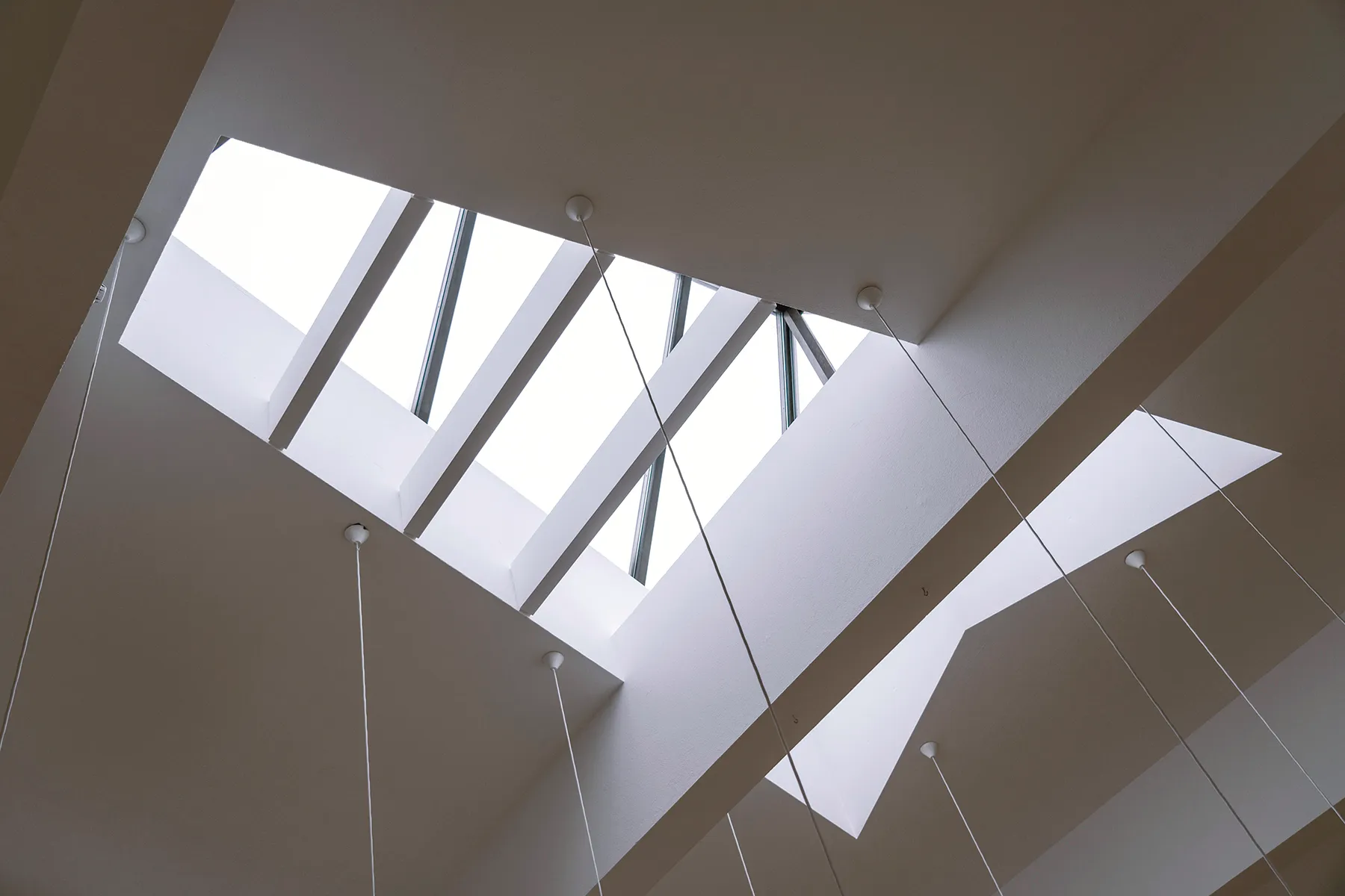 Angled white ceiling planes with rectangular skylights and thin hanging cables viewed from below inside the Nordic House