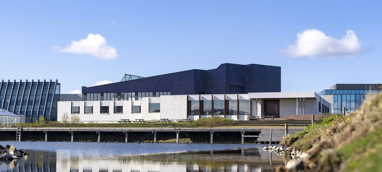 Wide exterior view of The Nordic House featuring a dark angular roof structure and white facade, with the building reflected in a calm pond in the foreground.